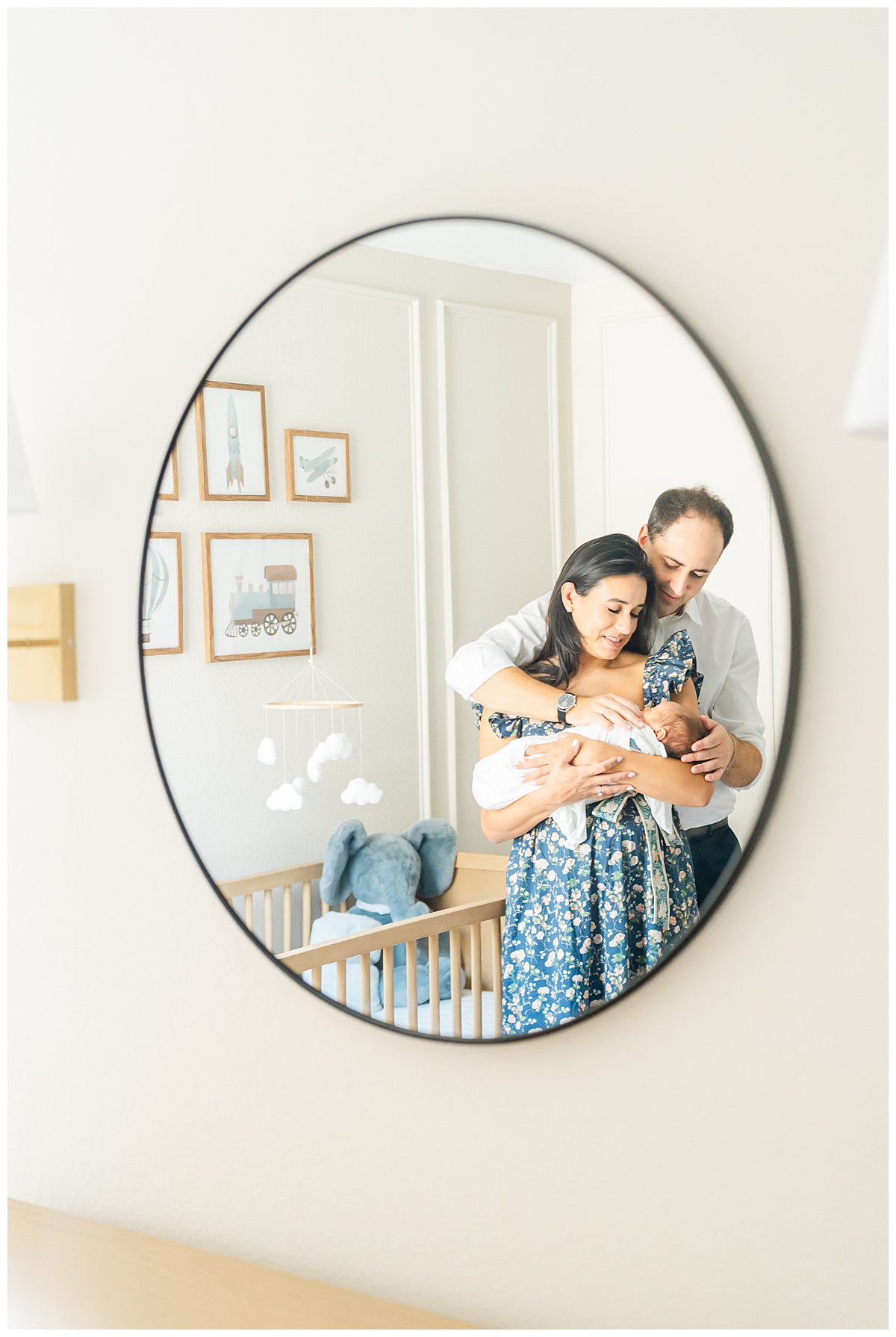 a mother and father stand in front of a crib in a nursery. The mother holds their newborn baby while her husband wraps his arms around them. The photo is captured by a Leander Photographer through the nursery mirror.