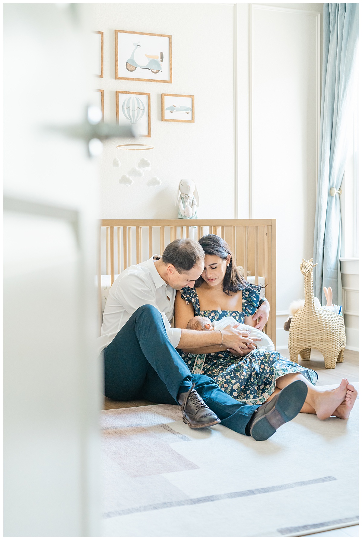 A mother and father sit in a nursery, in front of a crib. The mother holds their newborn baby. The parents look down at their baby while smiling, quietly captured behind the door by a Leander Photographer.