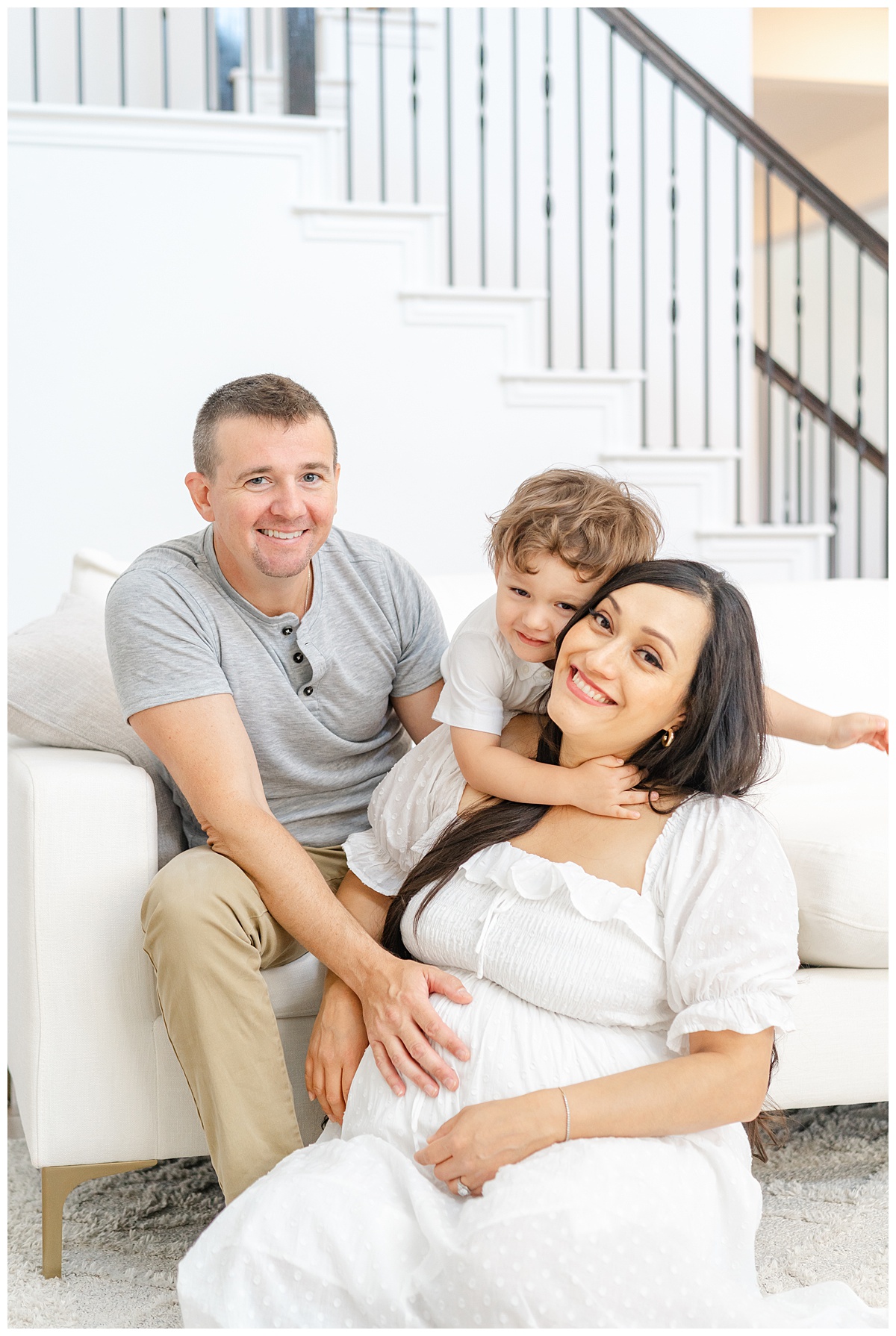 a family sits on a white couch. The pregnant mother sits on the floor while her toddler son wraps his arms around her neck, in this candid shot taken by a Leander Maternity Photographer.