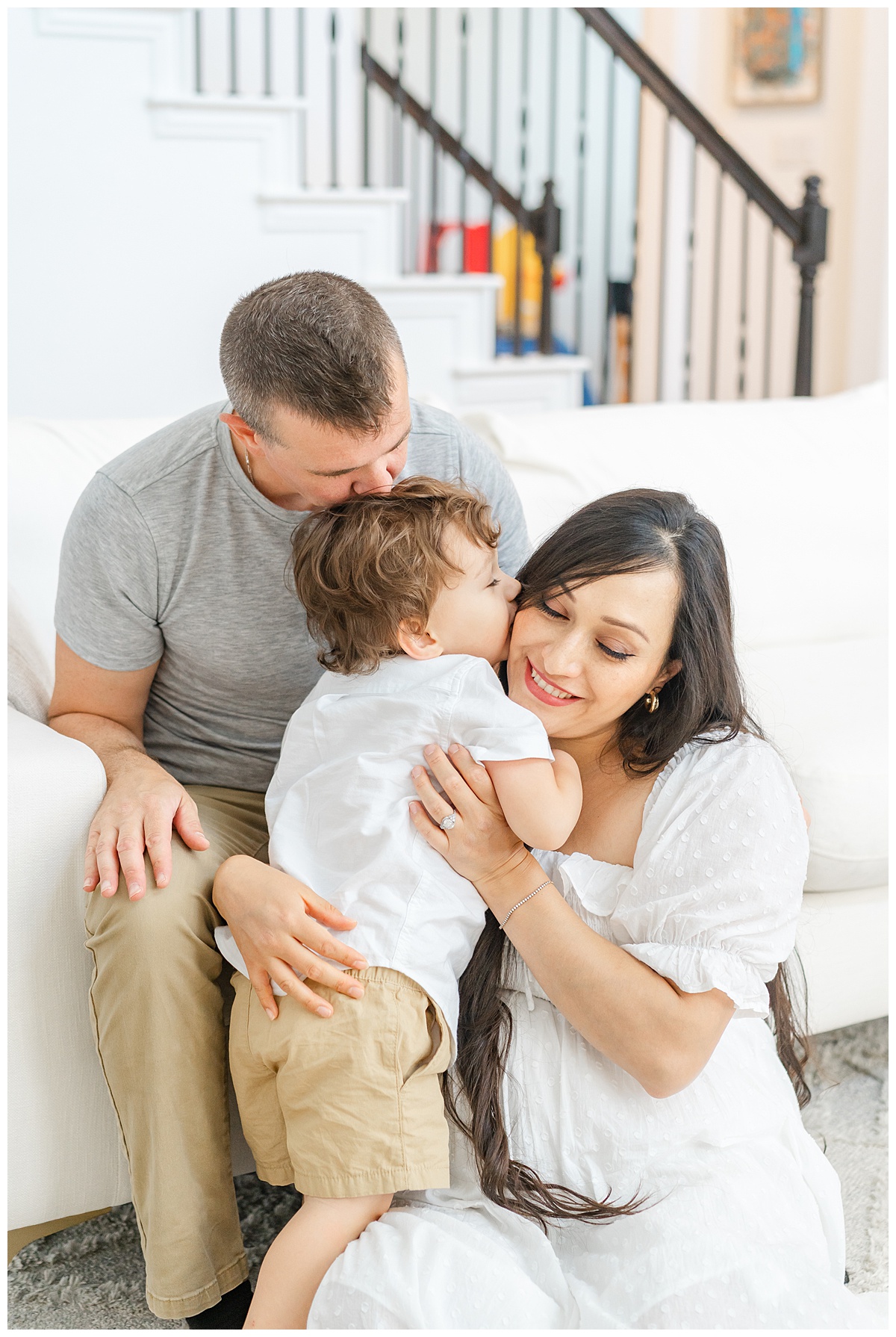A family of three kisses each other in front of their white couch, candidly captured by a Leander Maternity Photographer.