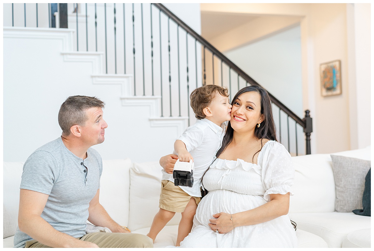 A family of three sits on a white couch. The toddler boy kissed his pregnant mother on the cheek in this candid shot captured by a Leander Maternity Photographer.