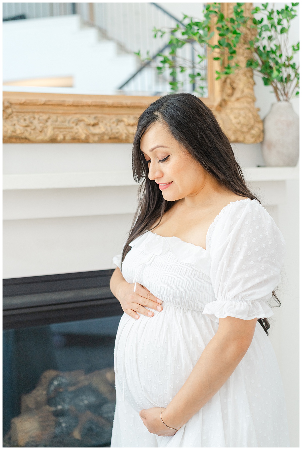 A pregnant mother dressed in a white dress stands in front of a fireplace in her Leander, TX home during her in-home maternity model call.