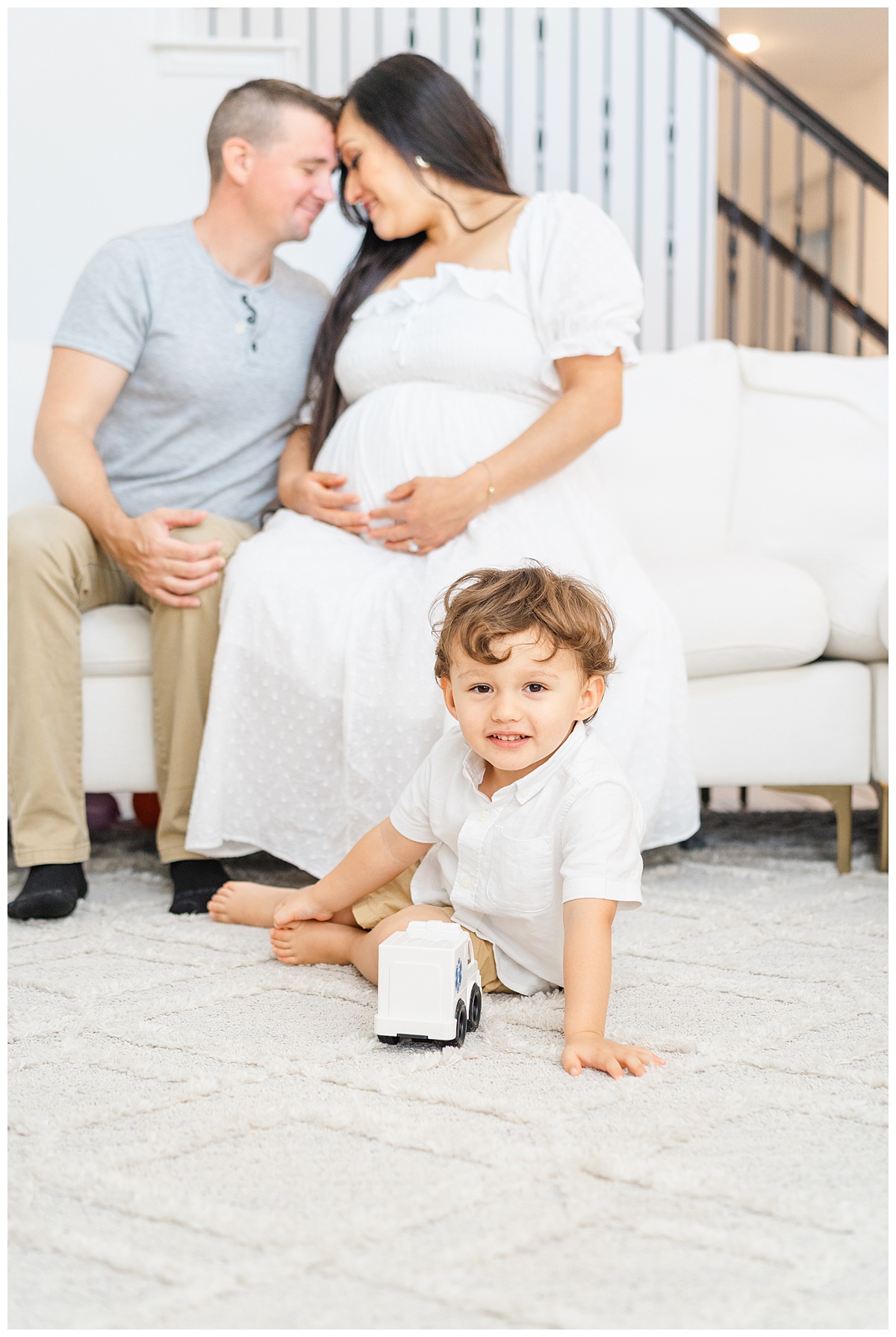 A toddler boy sits on the floor holding a toy truck. His parents sit on the couch behind him. The mother holds her pregnant belly during their in-home maternity model call in Leander, TX.