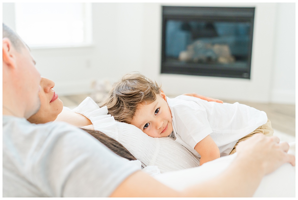 A toddler boy lays on top of his pregnant mother's belly. He looks at the camera of a Leander Maternity Photographer with a sweet smile.