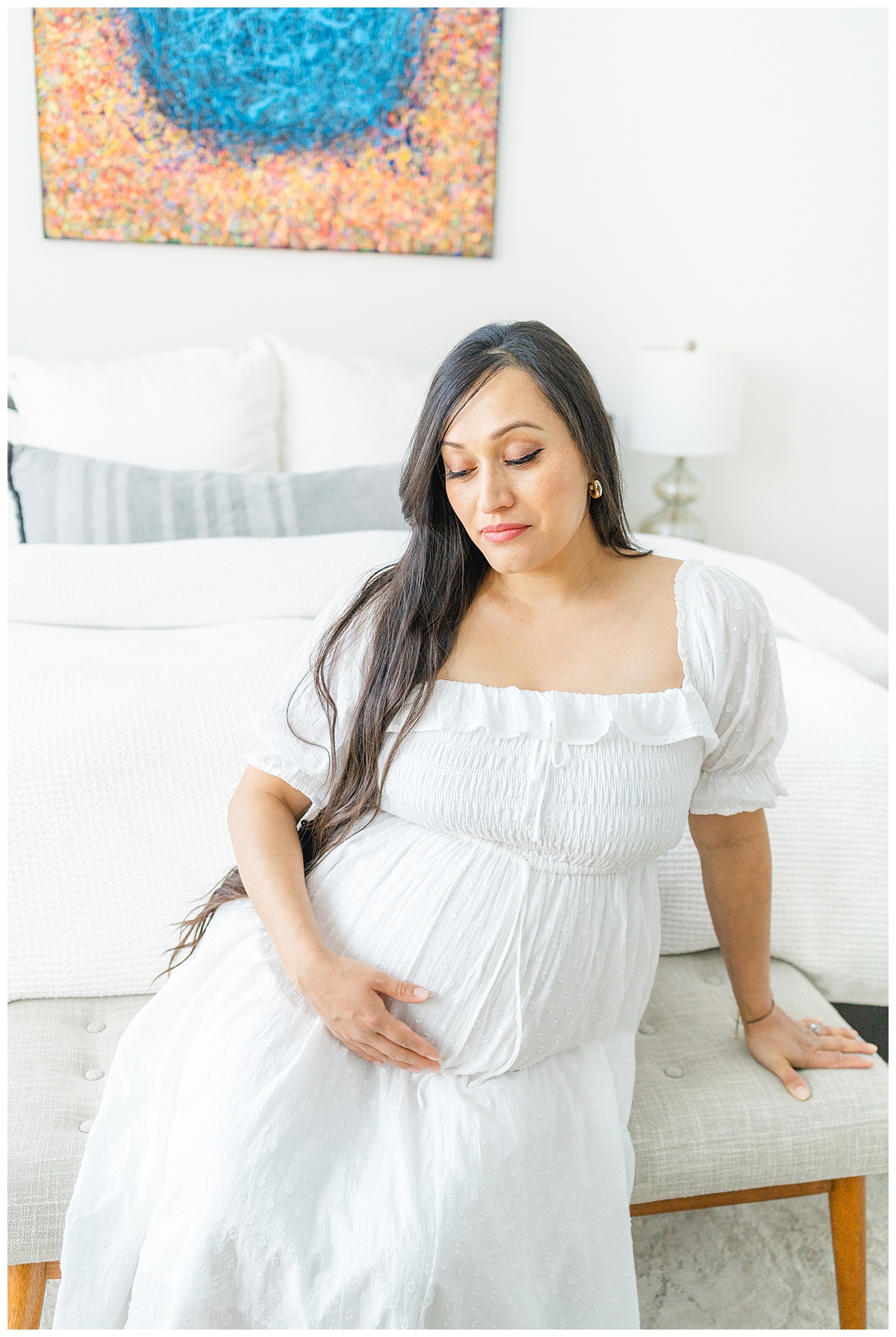 A pregnant mother in a white dress sits in front of her bed and looks down at her belly during her in-home maternity model call in Leander, TX.