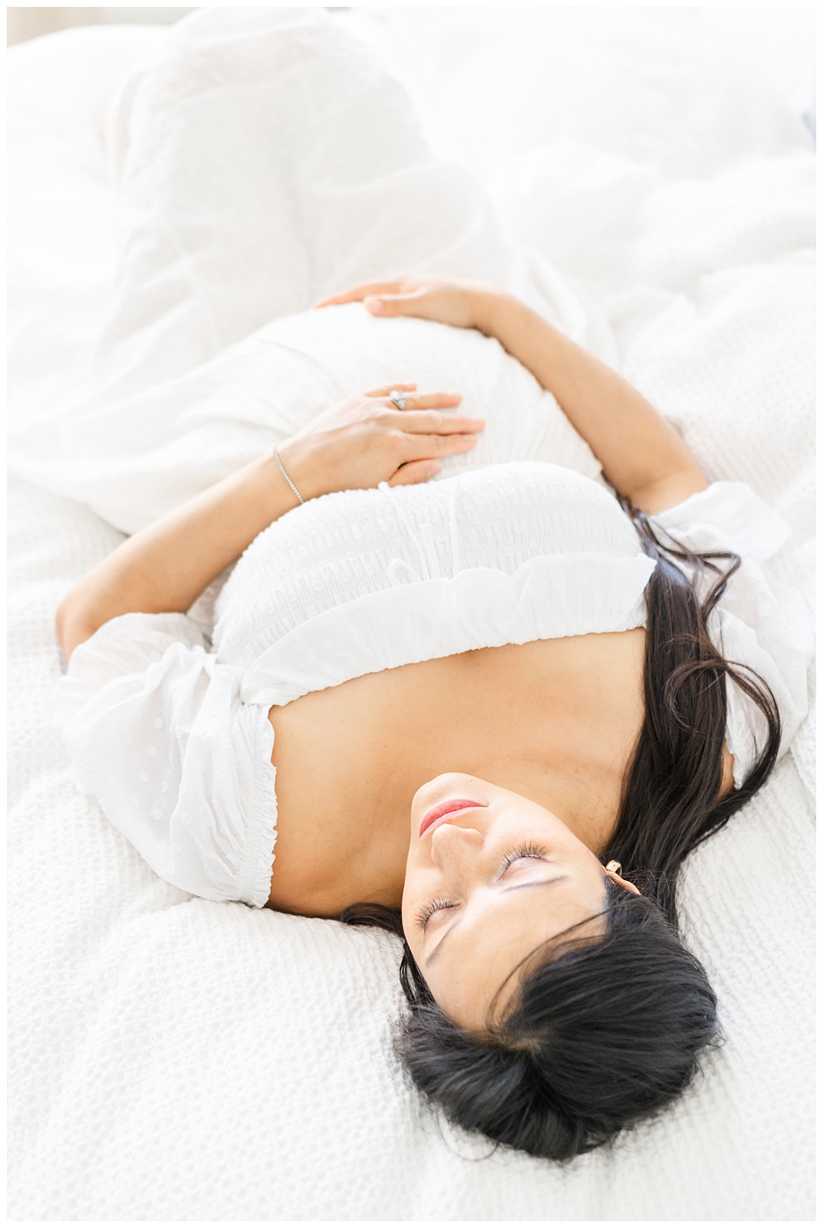 A pregnant woman in a white dress lays on a white bed. She holds her pregnant belly with both hands during her in-home maternity session in Leander, TX.