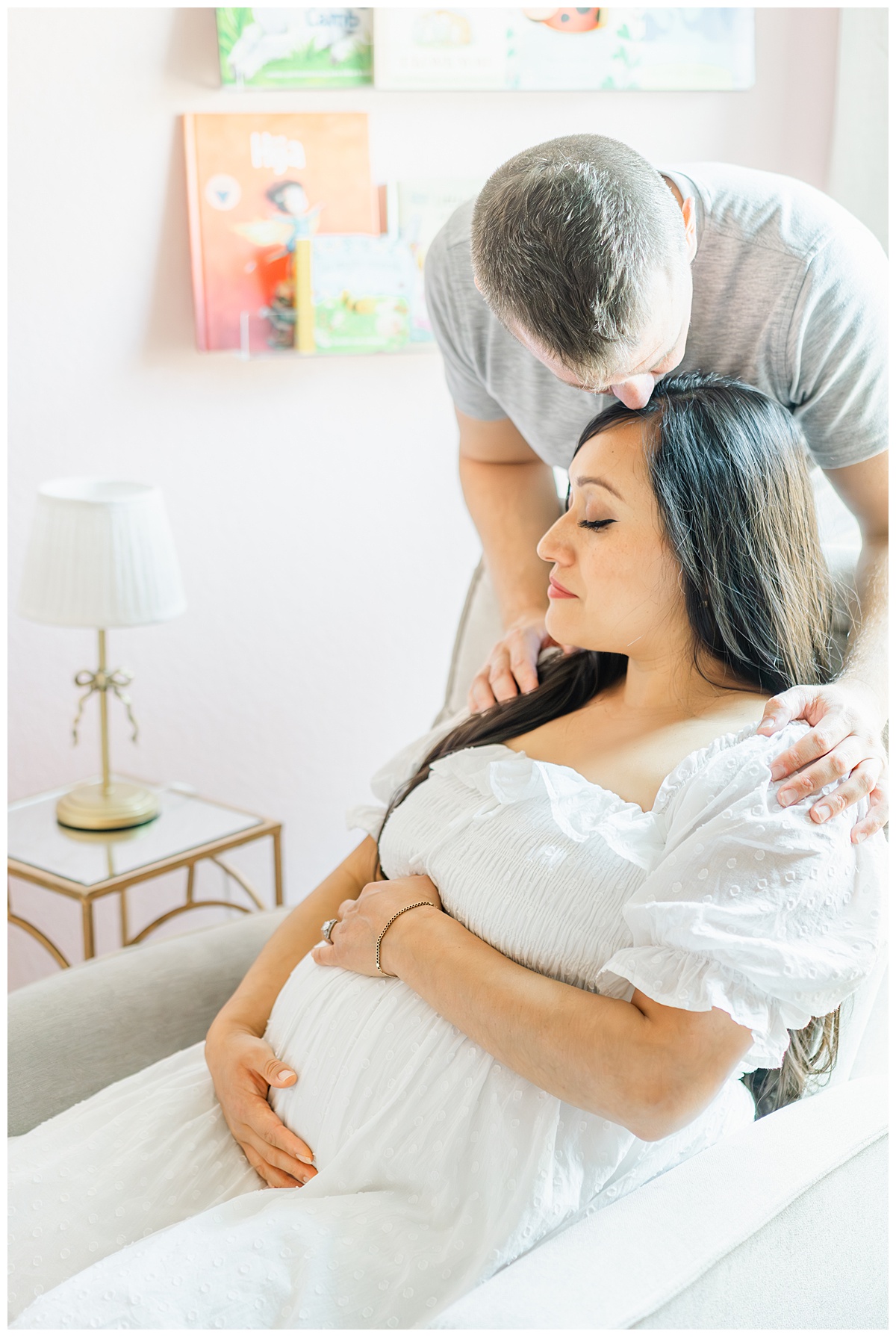a pregnant mother sits in a rocking chair in her baby girl's nursery. Her husband stands behind her. He bends down to kiss her on the forehead during their in-home maternity model call in Leander, TX.