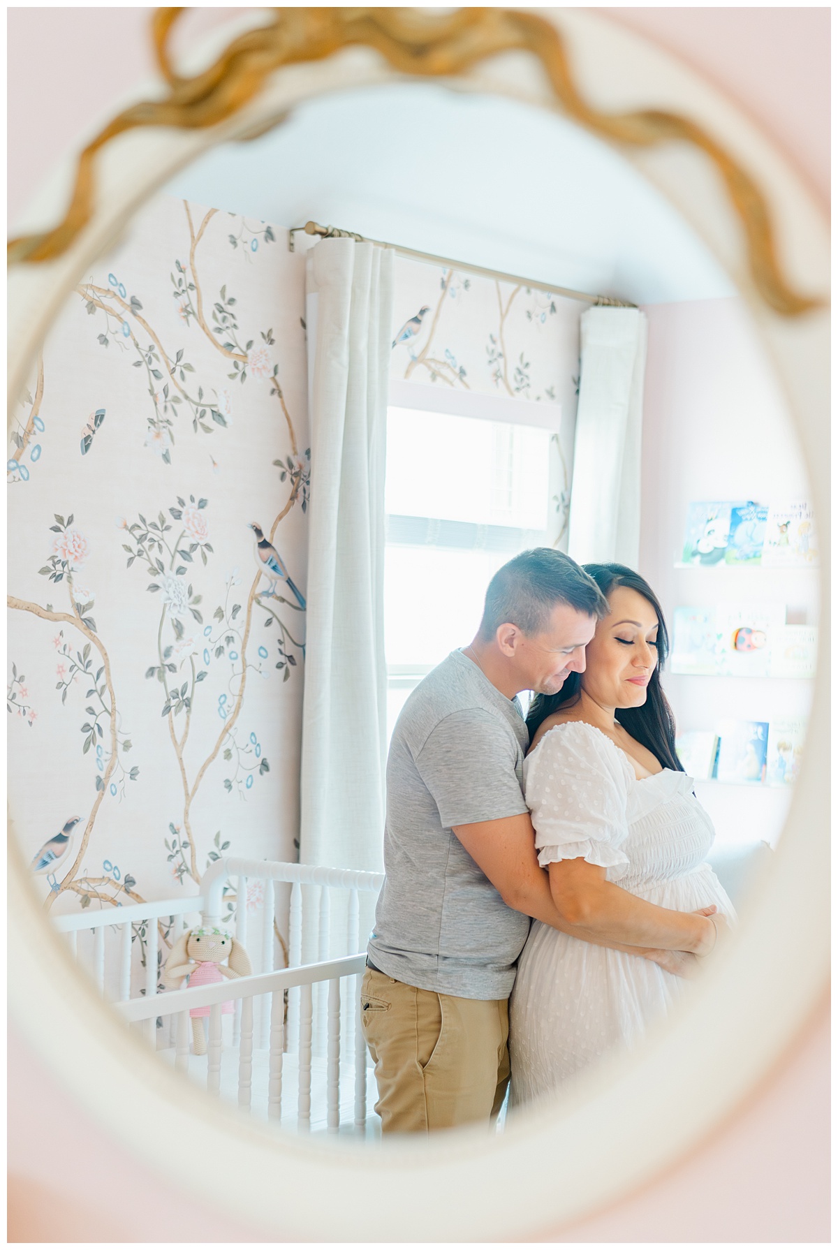 A couple is captured by a Leander Maternity Photographer through an oval mirror with a golden bow. The woman is pregnant and holds her pregnant belly. Her husband stands behind her and wraps his arms around her belly.