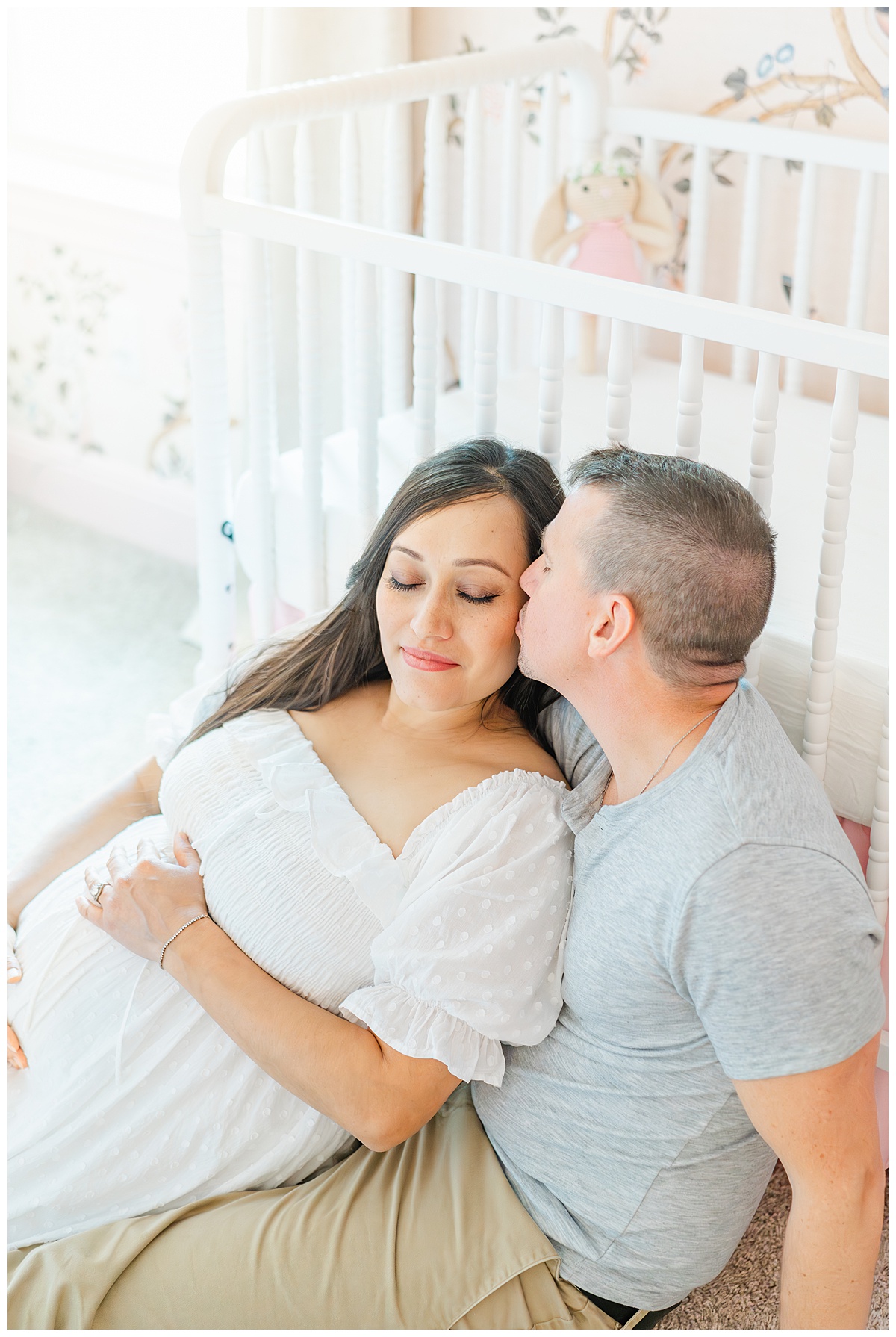 A couple sits in front of a white crib in a nursery. The pregnant woman leans her back against her husband while he kisses her on the cheek during their in-home maternity session in Leander, TX.
