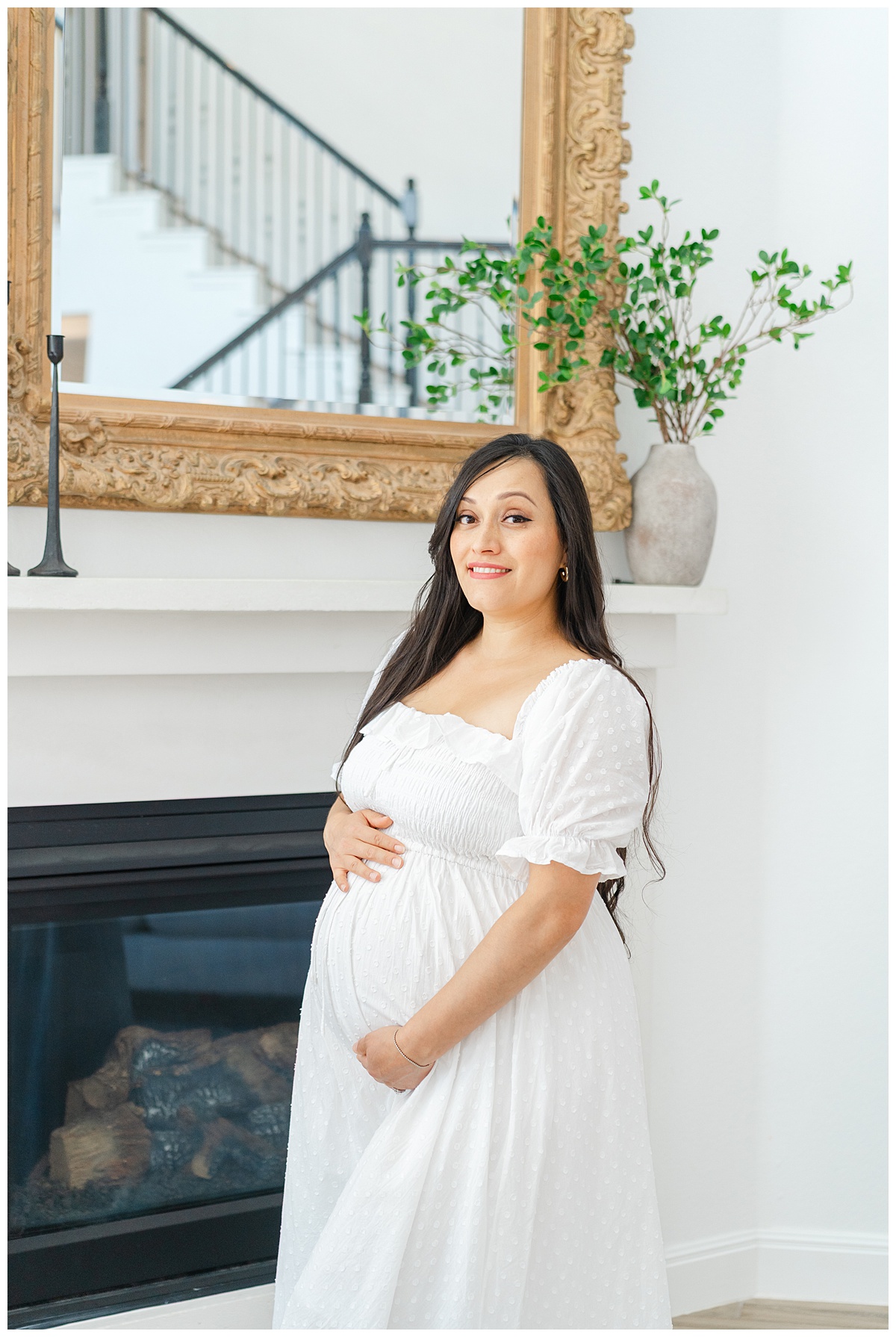 A pregnant woman dressed in a white dress stands in front of a fireplace while smiling at the camera of a Leander Maternity Photographer.