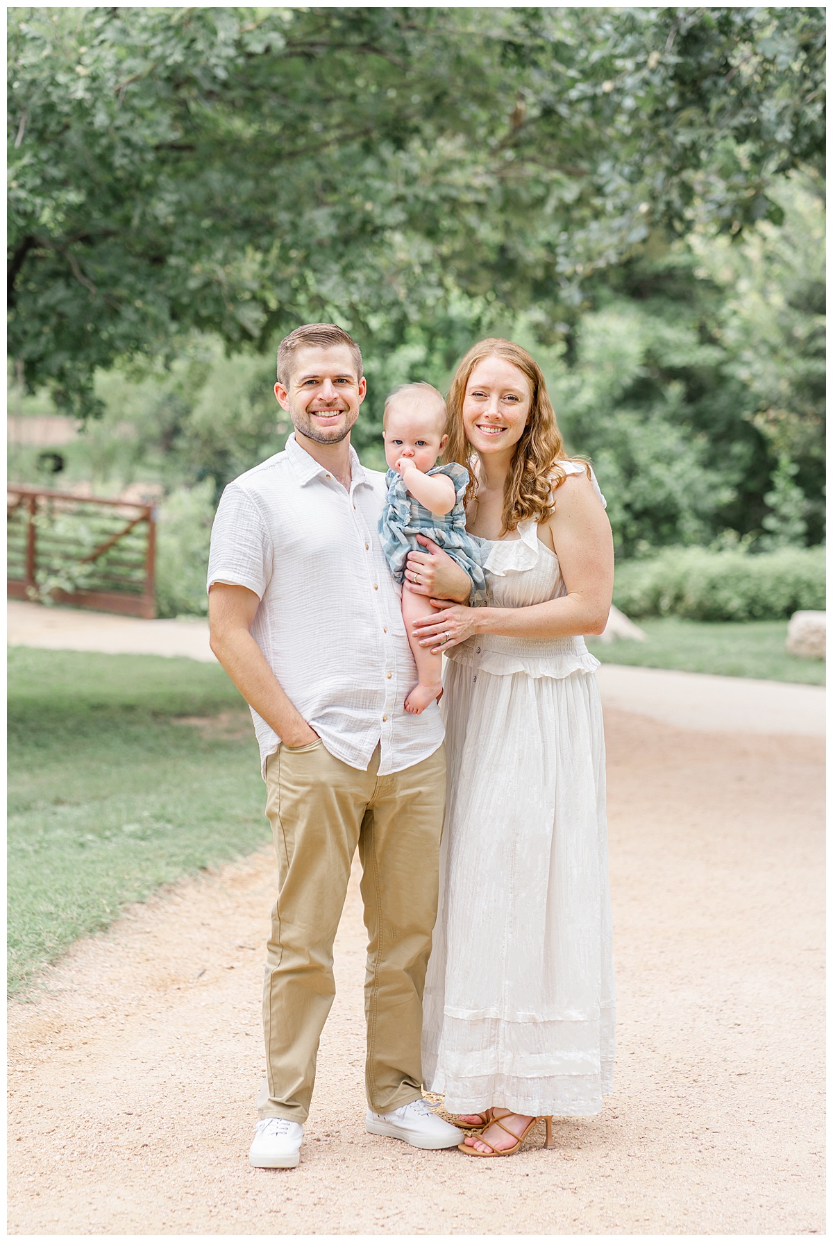 A family of 3 stand on a path in front of a bridge at Katherine Fleischer Park. The father holds the baby girl in between him and his wife. They all look at the camera. The baby hads her hand in her mouth. Her parents smile. 