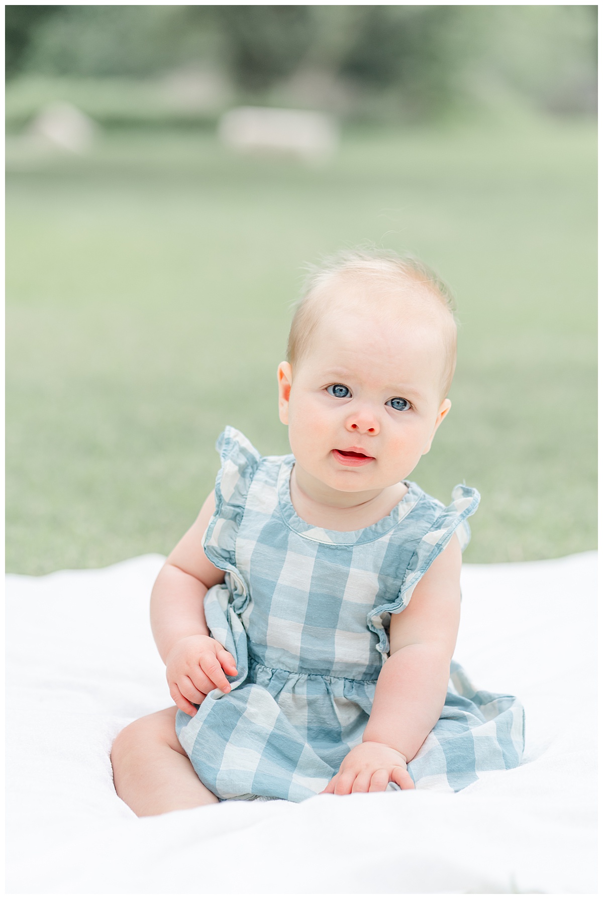 Austin Photographer | A baby girl dressed in a blue gingham dress sits on a white blanket on a grass area at Katherine Fleischer Park. 
