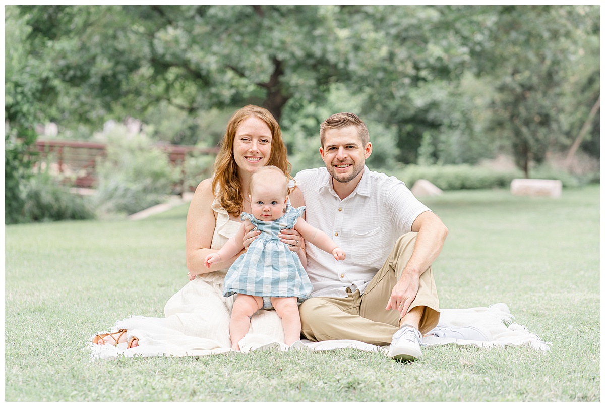 a family of three sits on a white blanket at Katherine Fleischer Park in front of a bridge. The mother holds their baby girl. They all look at the camera and smile. 