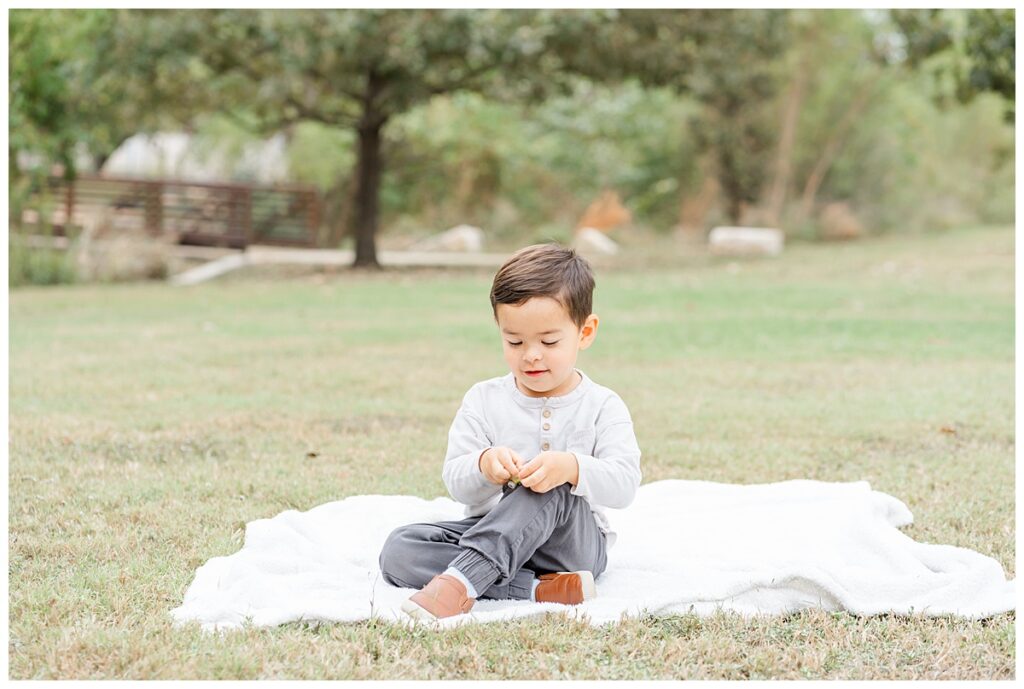 a toddler boy dressed in a light grey shirt and dark grey pants sits on a white blanker at a park and plays with something in his hands.