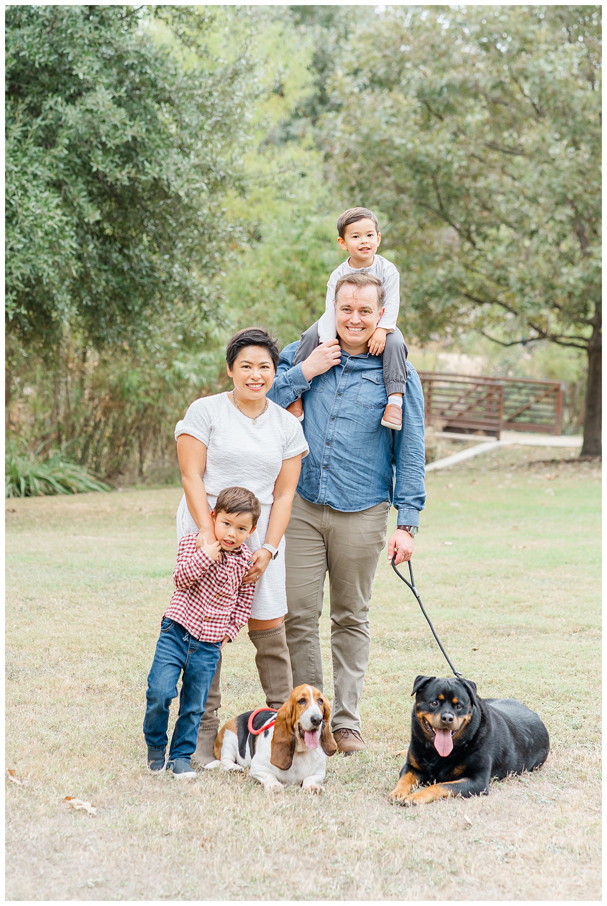 A family stands on the grass at an Austin park with their two dogs. The father holds the younger boy on his shoulders while the older boy stands in front of his mother and holds her hand.