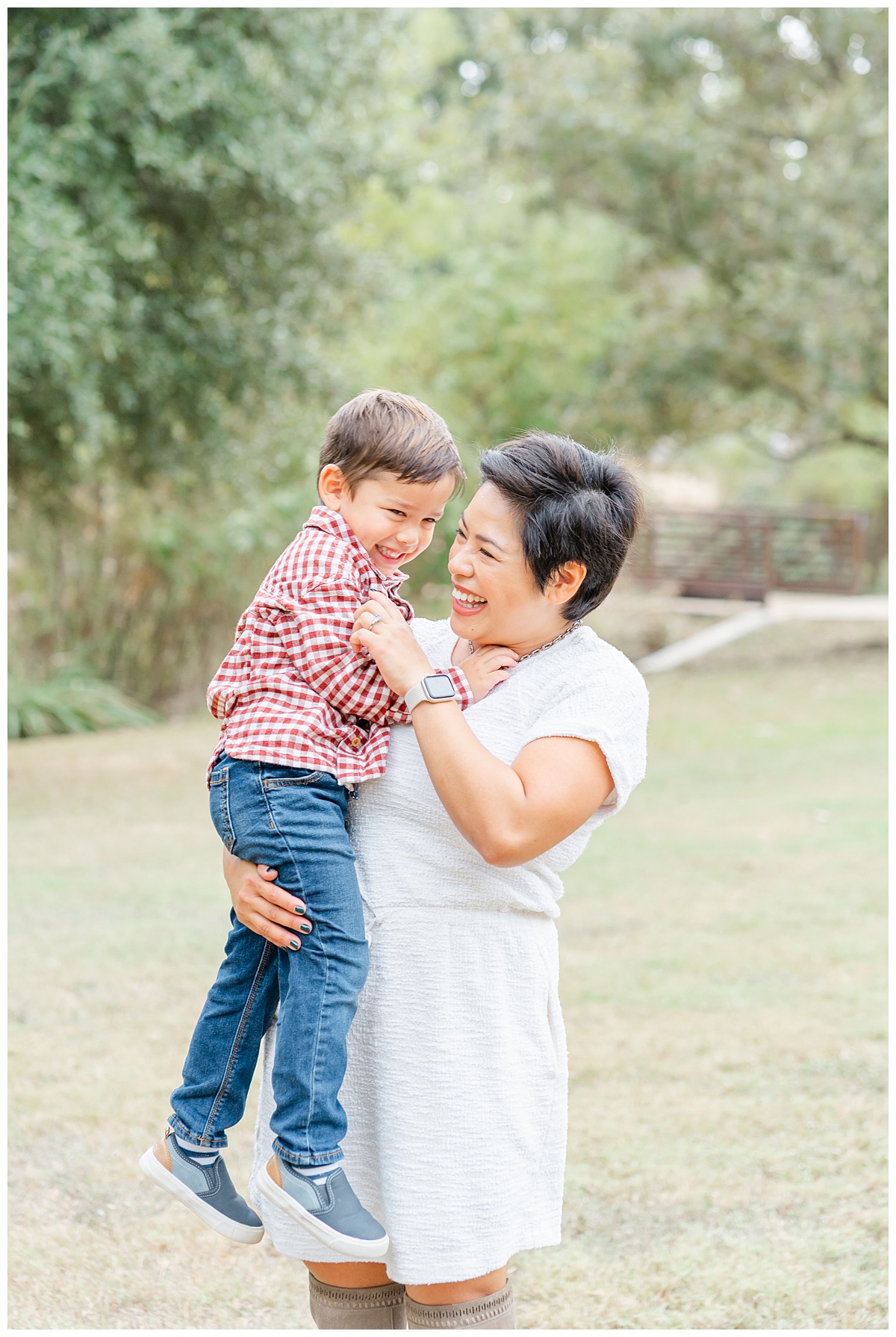 a mother dressed in a white dress stands and holds her young son while tickling him.