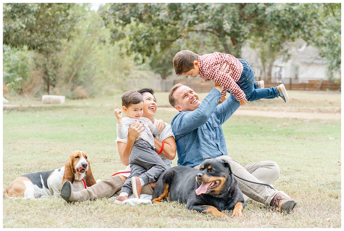a family sits on a white blanket at an Austin park. The father holds up one of their two young boys up in the air while the mother and younger boy look at him and smile. Their two dogs lay in front of them.