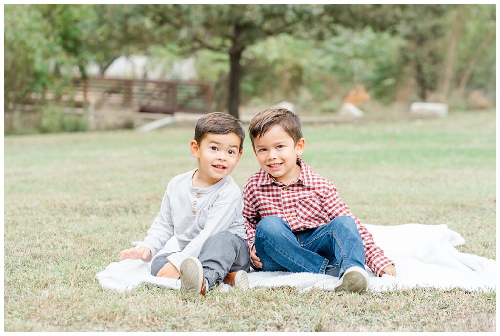 two toddler boys sit on a white blanket and pose for their family photos at an Austin park.