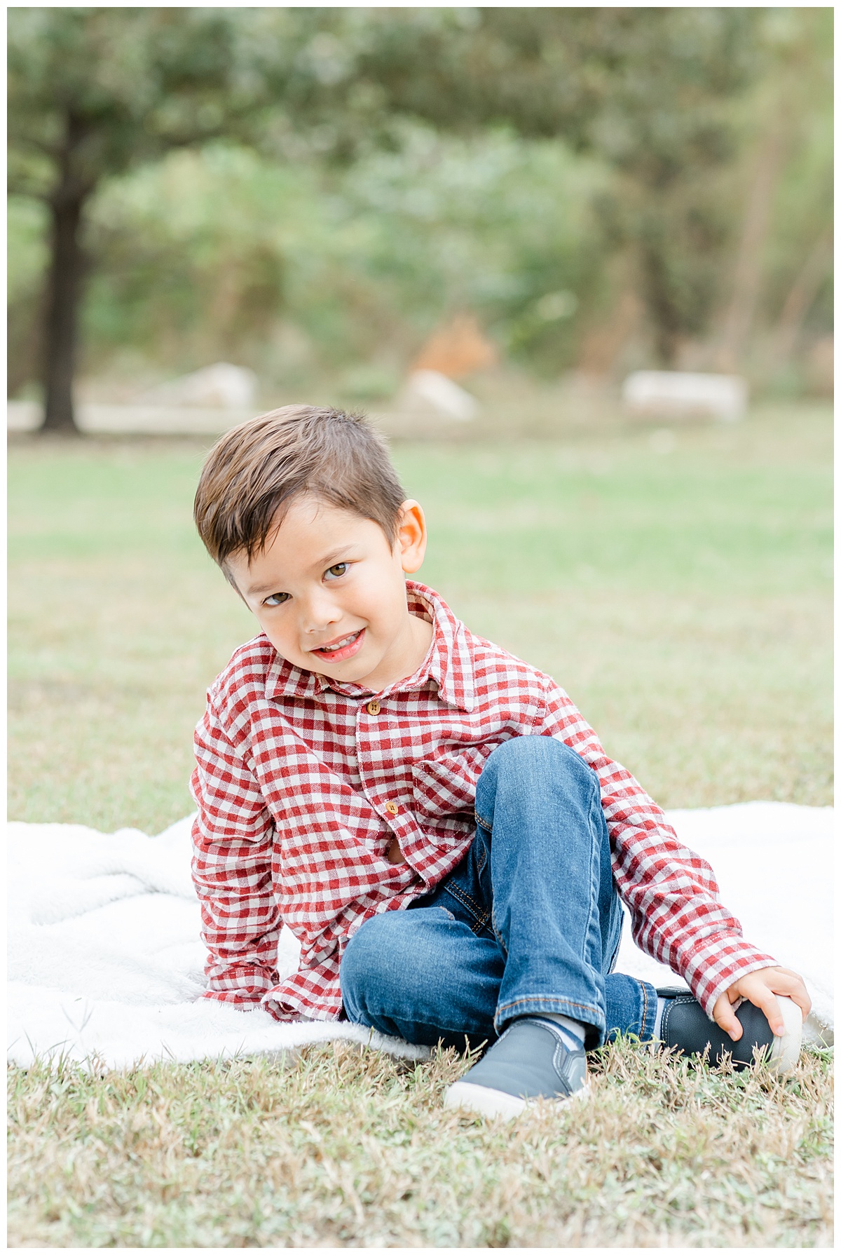 a young boy dressed in a red plaid shirt and dark blue jeans sits on a white blanket at a park and looks at his Austin family photographer.