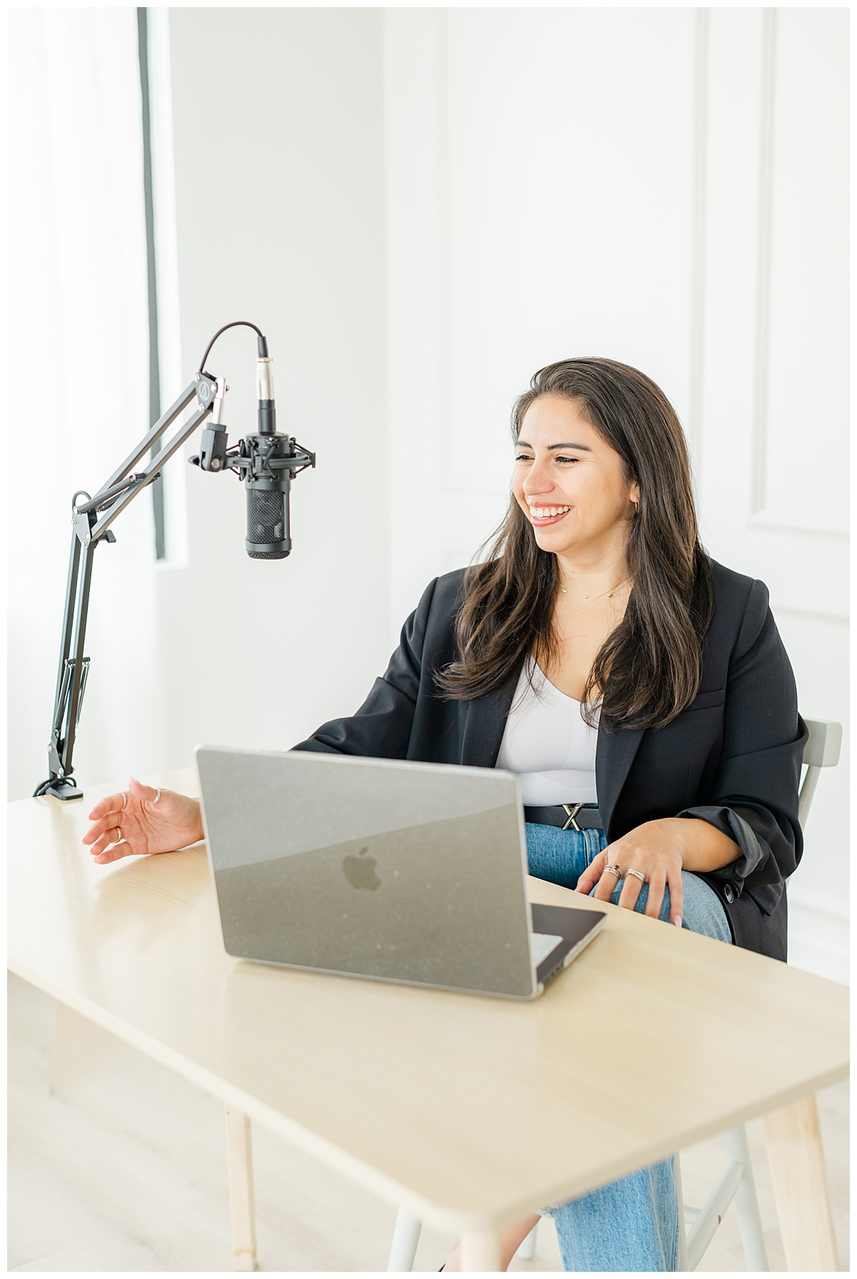 a woman sits at a desk in a white photo studio and laughs while speaking into a microphone. 