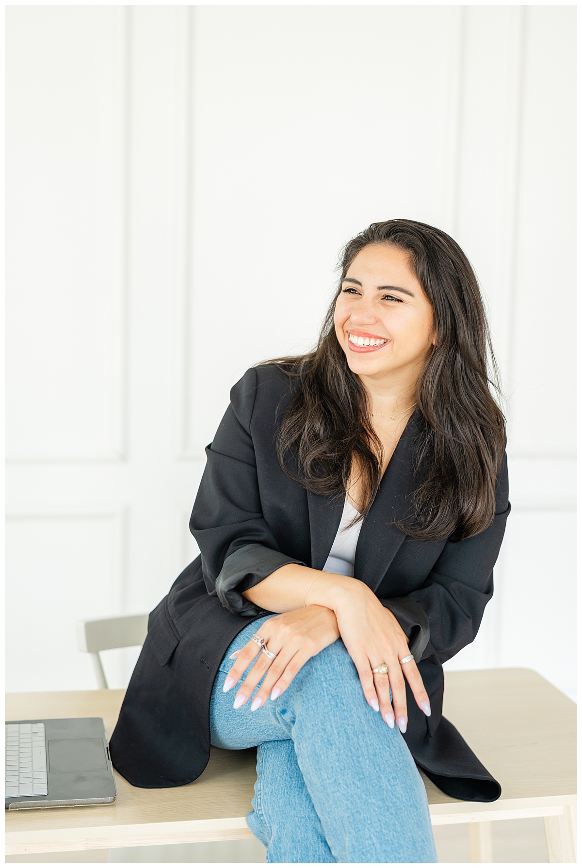 a woman with long dark hair wearing a black blazer sits on a desk and smiles during her branding photo session. 