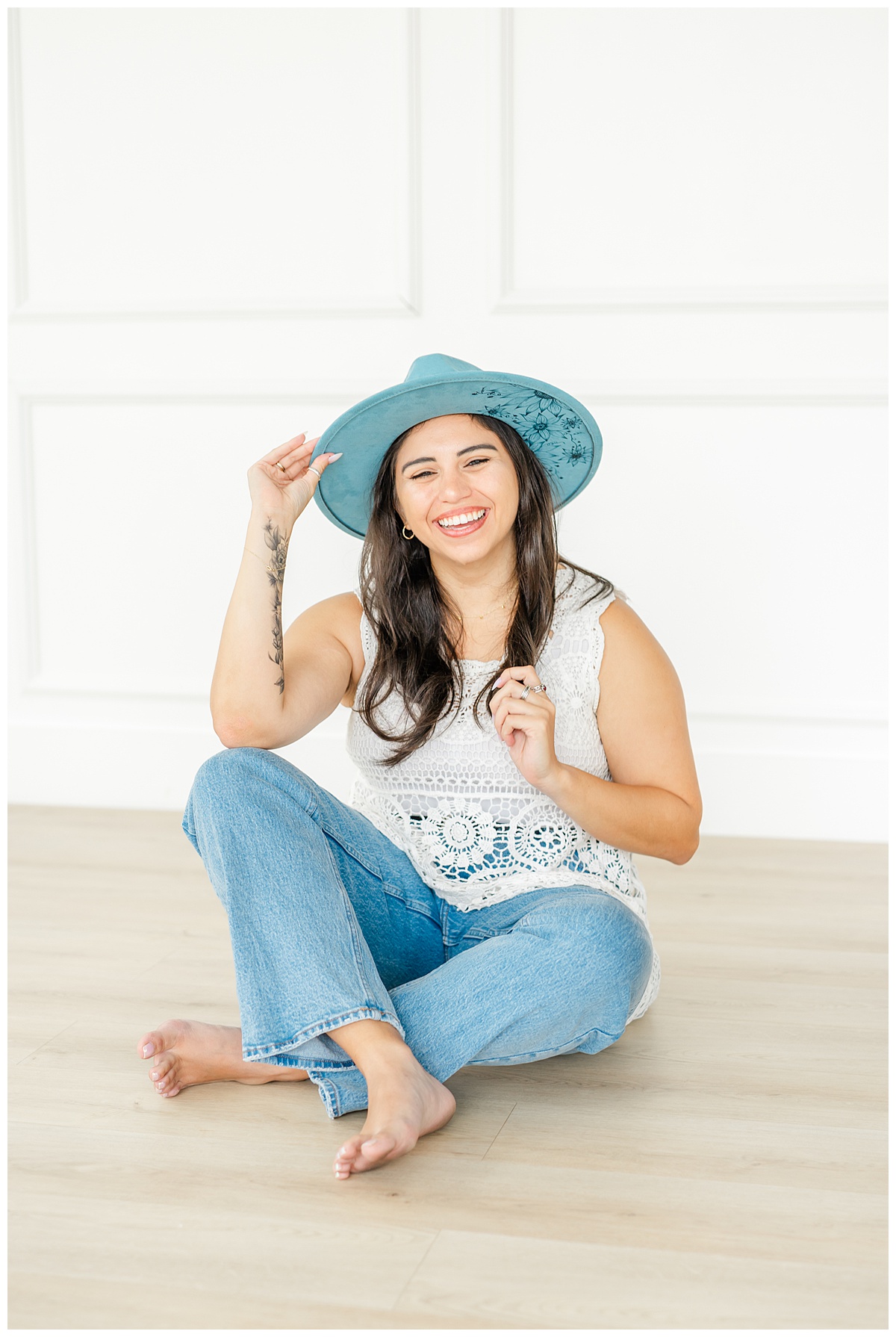 a woman sits on the floor of an Austin photo studio and holds onto her blue hat while smiling. 