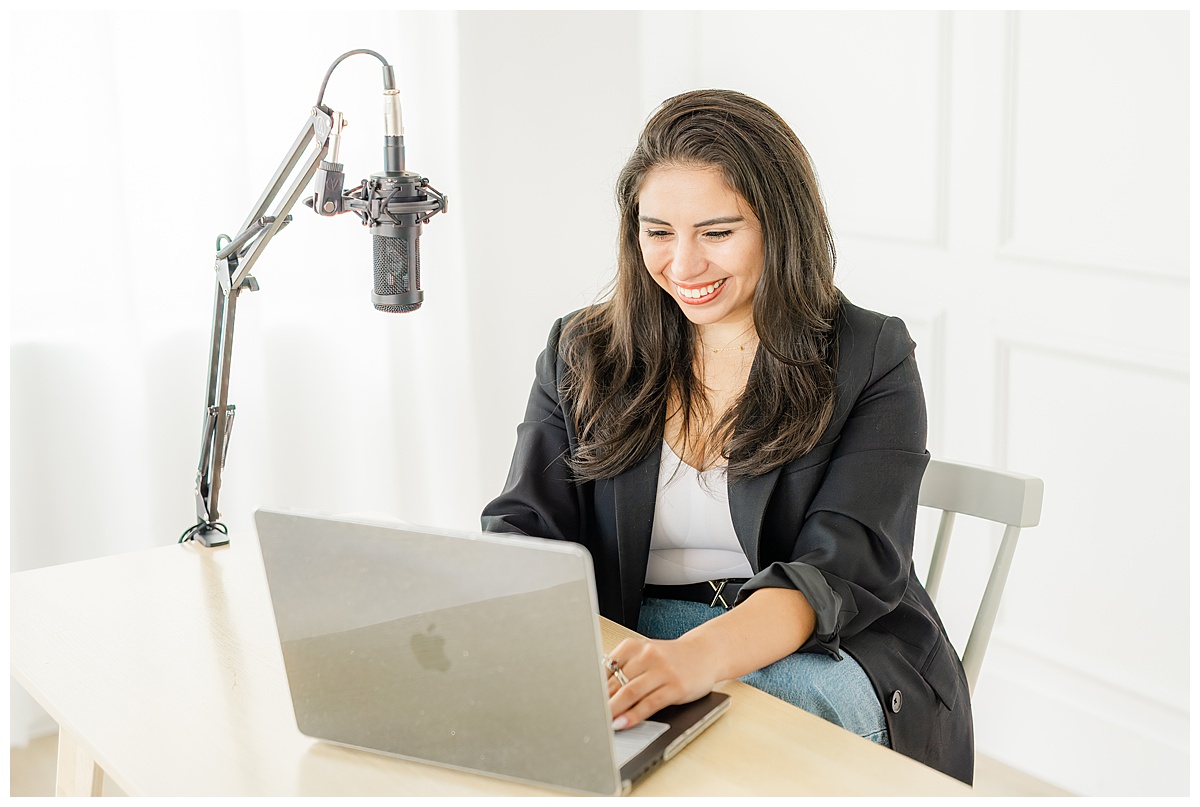 an Austin podcast host sits at a table in a white studio and types on her laptop. 