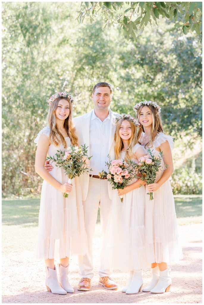 A father dressed in an ivory suit stands with his three teenage daughters at an Austin park. They each hold a bouquet, looking like a wedding party. 