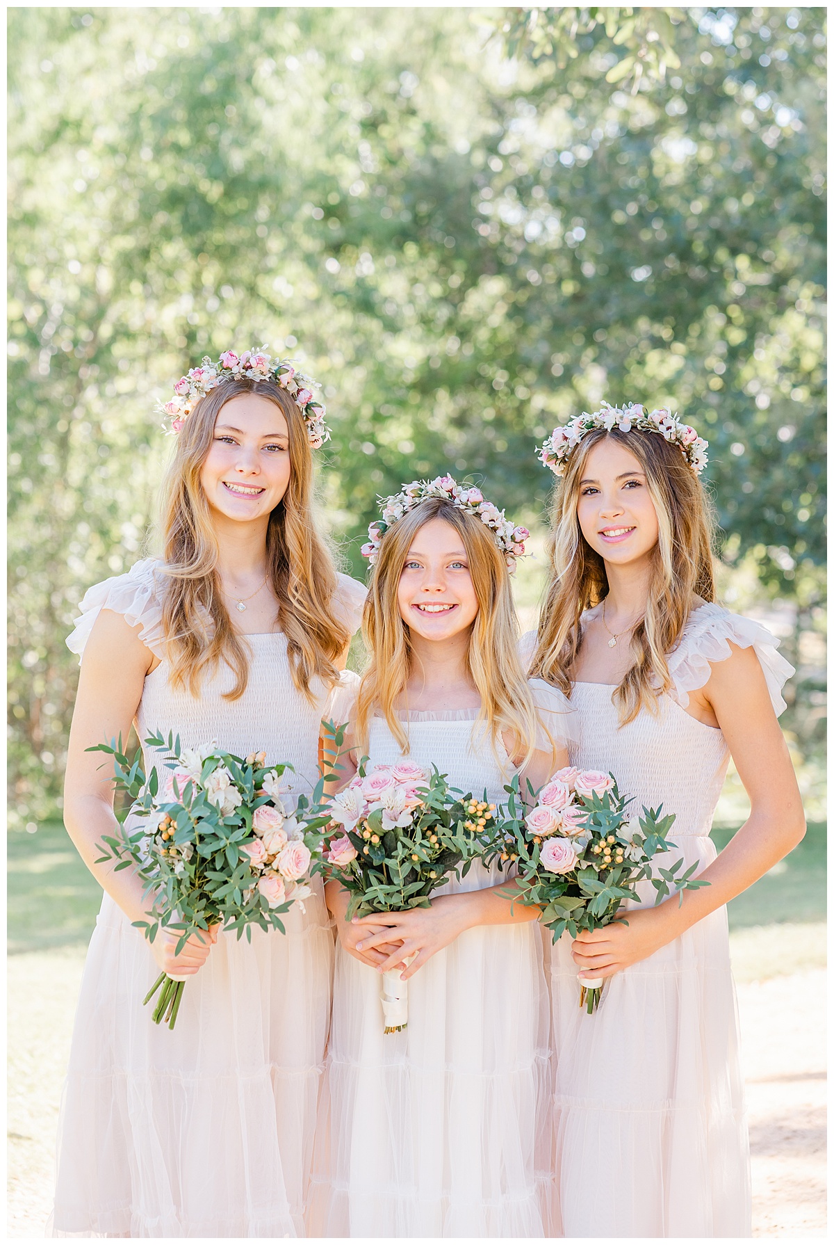 Three teenage girls wearing matching ivory dresses and floral crowns stand together while smiling at their Austin family photographer.