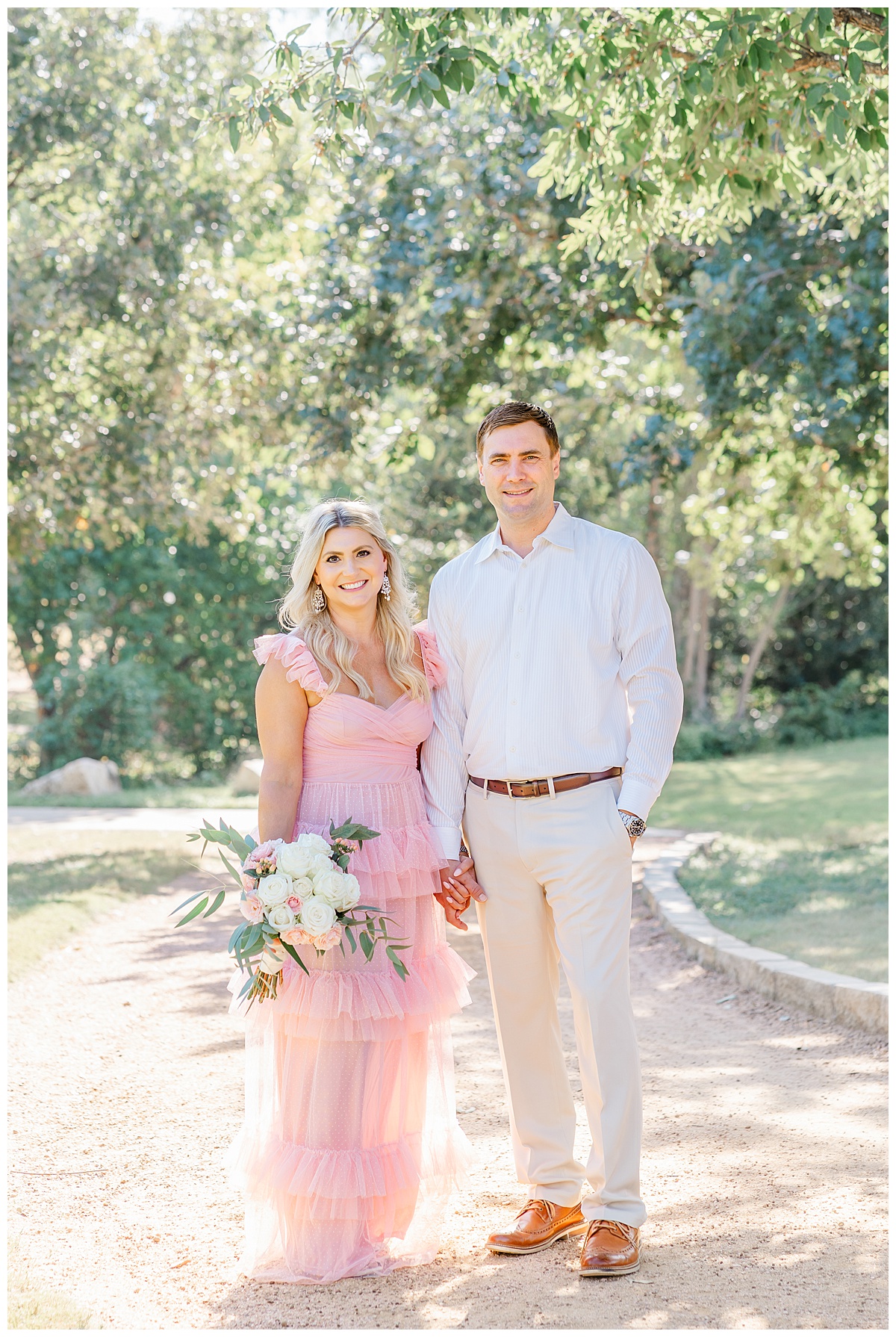 A couple with a woman dressed in a long pink dress and a man dressed in an ivory suit stand under a tree holding hands and smile at their Austin family photographer.