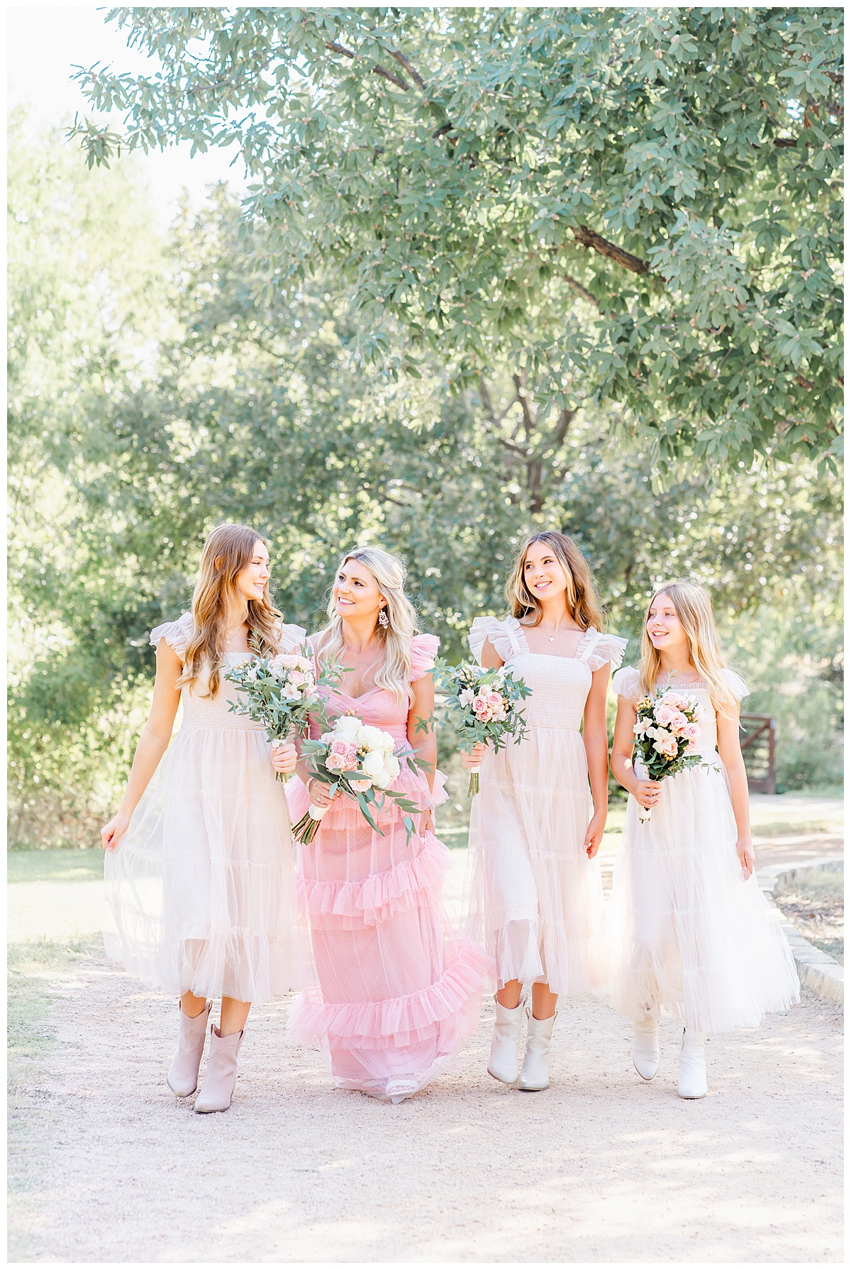 A mother dressed in a long pink dress walks up a path at an Austin park with her three teenage daughters. They each hold a bouquet to celebrate their anniversary with family photos.