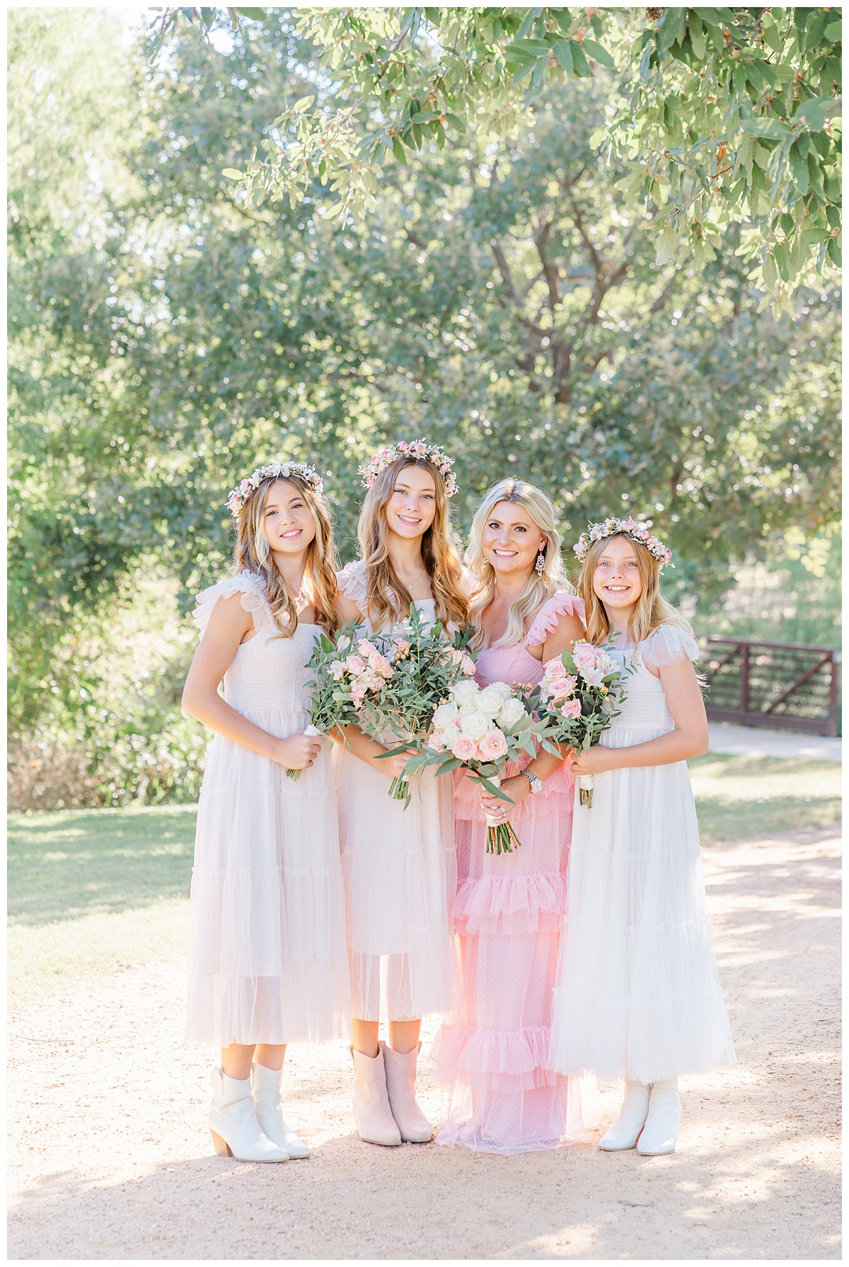 a mother and her three teenage daughters stand under a tree at an Austin park. The mom is dressed in a long pink dress while her daughters wear matching ivory dresses. They each hold a bouquet for anniversary photos.