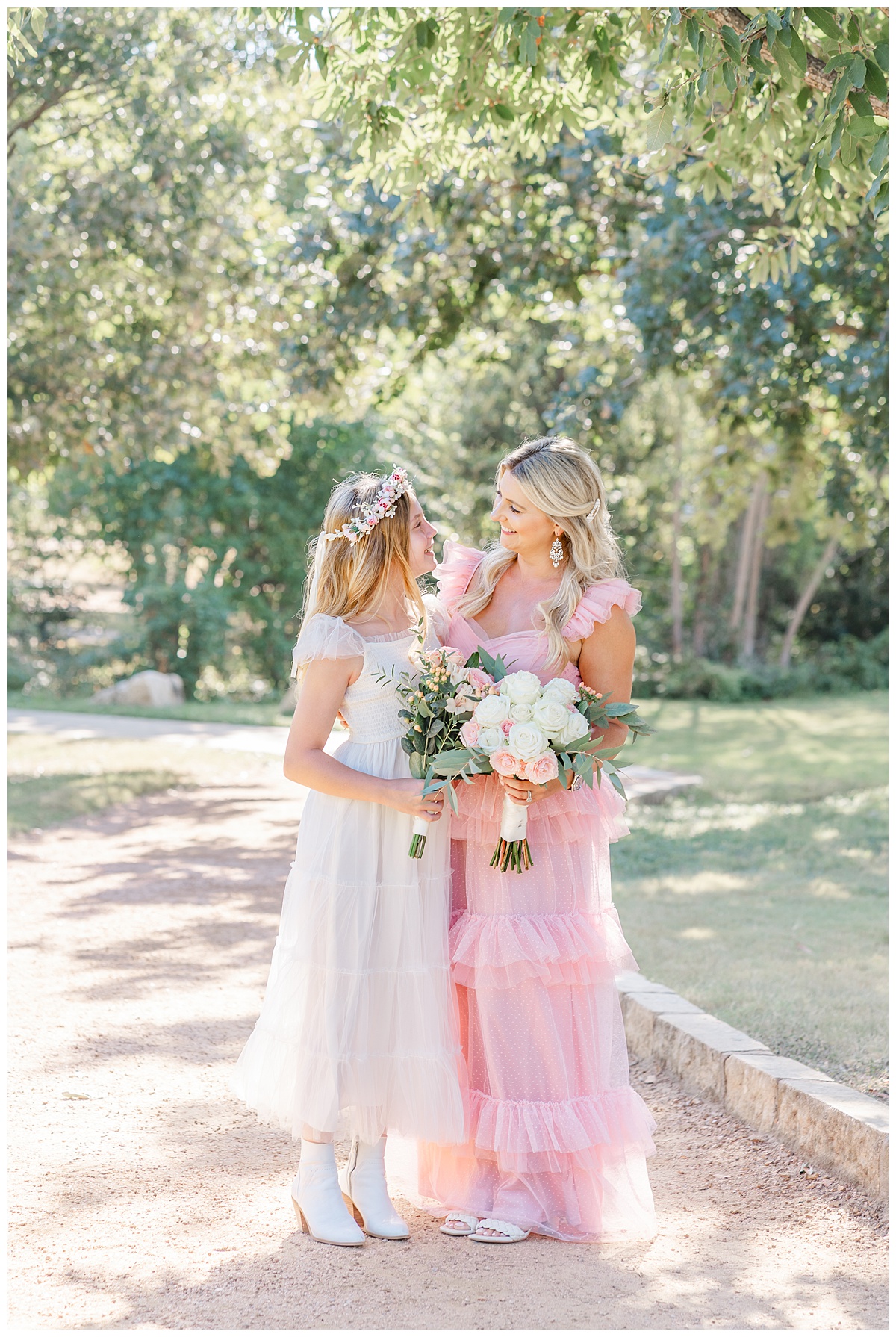 A mother dressed in a long pink tulle dress looks at her teenage daughter with a smile. They stand together each holding a bouquet. 