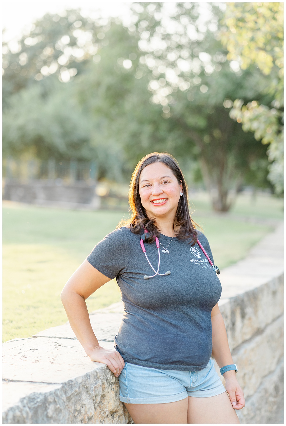 a mama coach and registered nurse stands against a pony wall at Butler Metro Park for her headshots. She wears a grey shirt and denim shorts, and has a stethoscope around her neck.