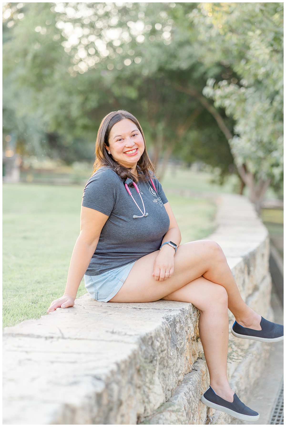 a registered nurse with a stethoscope around her neck sits on the edge of a pony wall at Butler Metro Park in Austin, TX and smiles at the camera of an Austin family and newborn photographer.