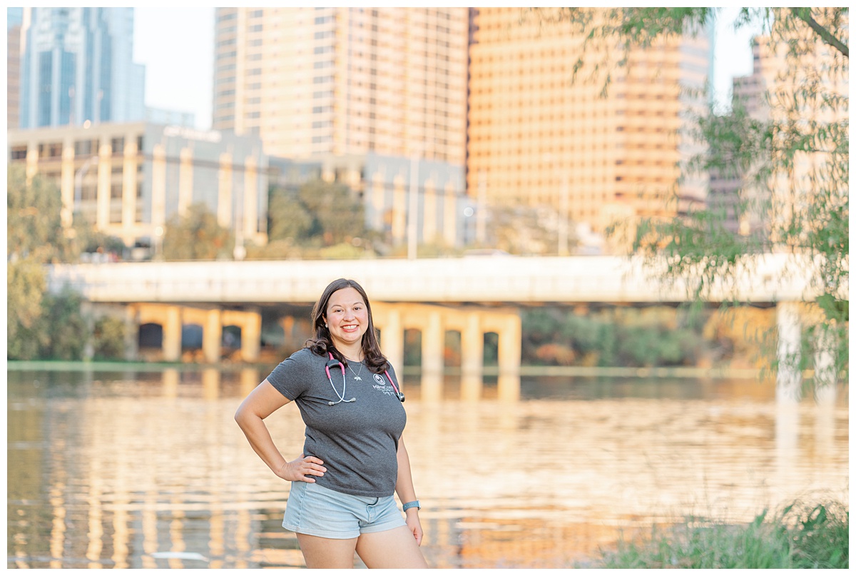 a mama coach and nurse stands in front of the Austin town lake and the downtown skyline. She wears a pink stethoscope around her neck.