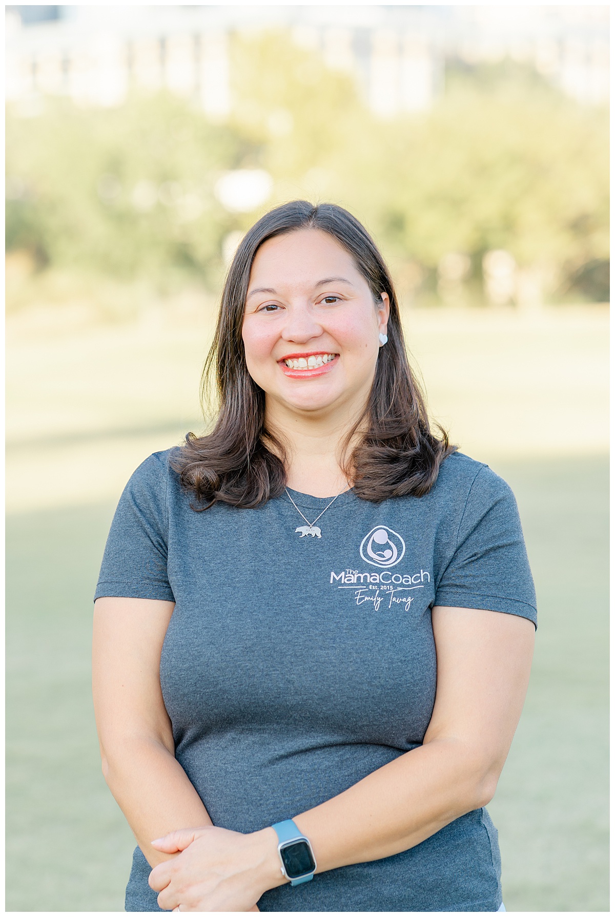 a woman wearing a grey shirt with a logo spelling Mama Coach stands at an Austin park for her headshot session.