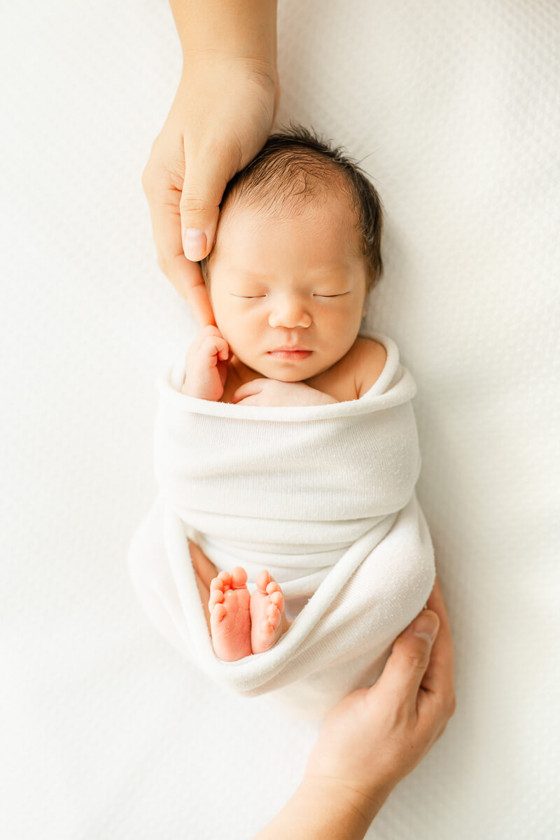 newborn baby sleeps on white surface and parents hold her hands and feet.