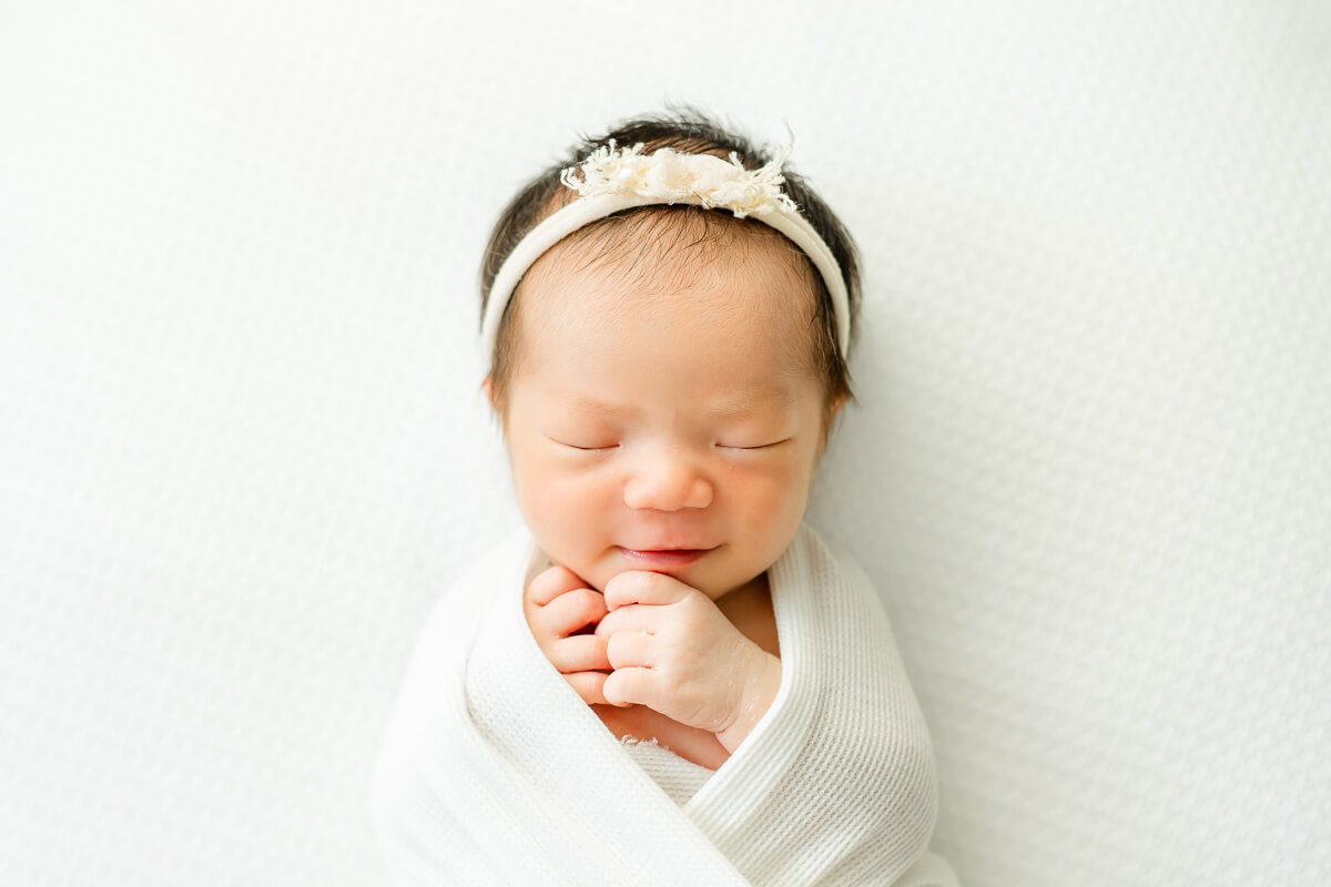 smiling infant wrapped in white blanket with floral headband sleeps on white surface while captured by an Austin newborn photographer.