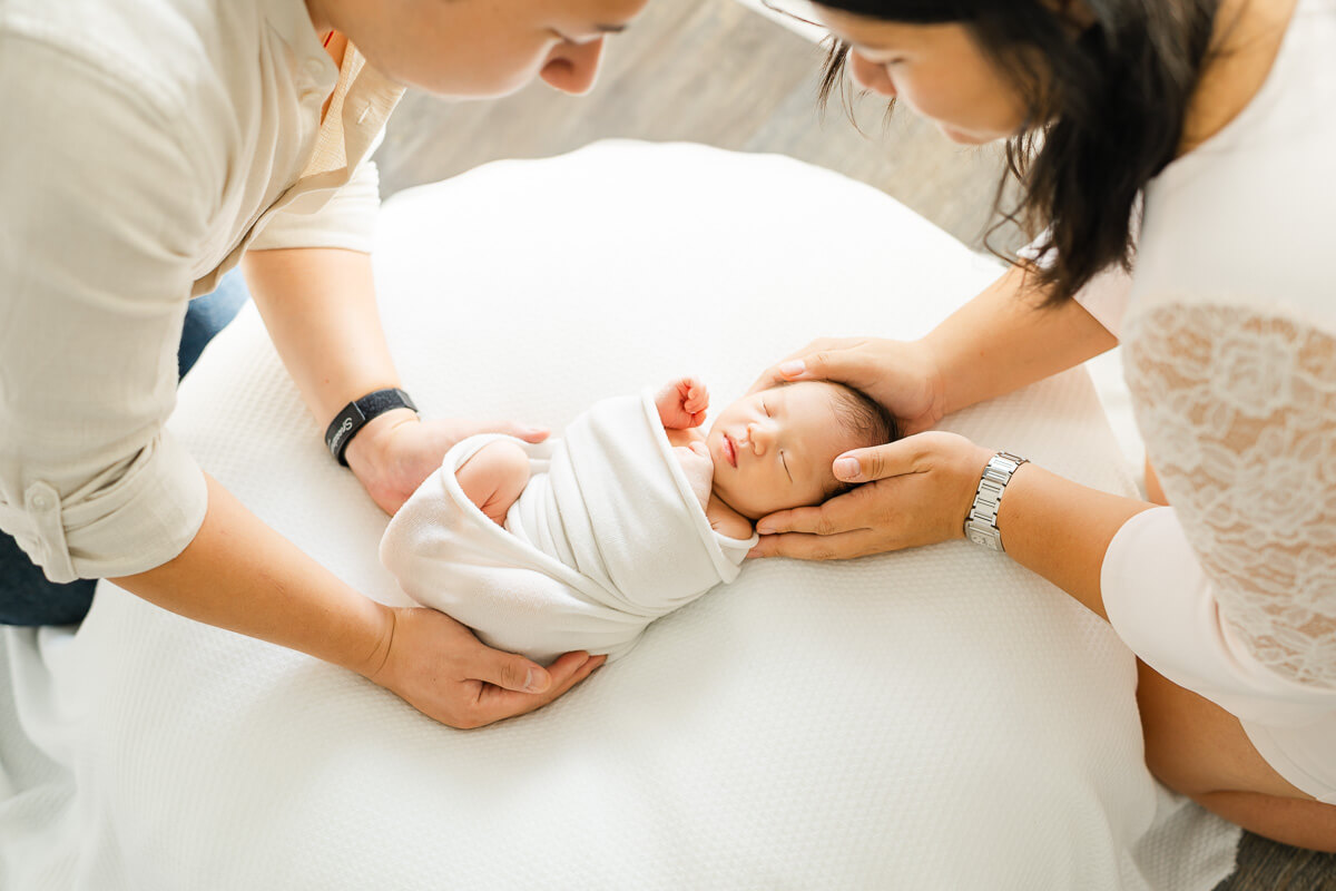 parents hold baby's head and feet while she sleeps on white bean bag.