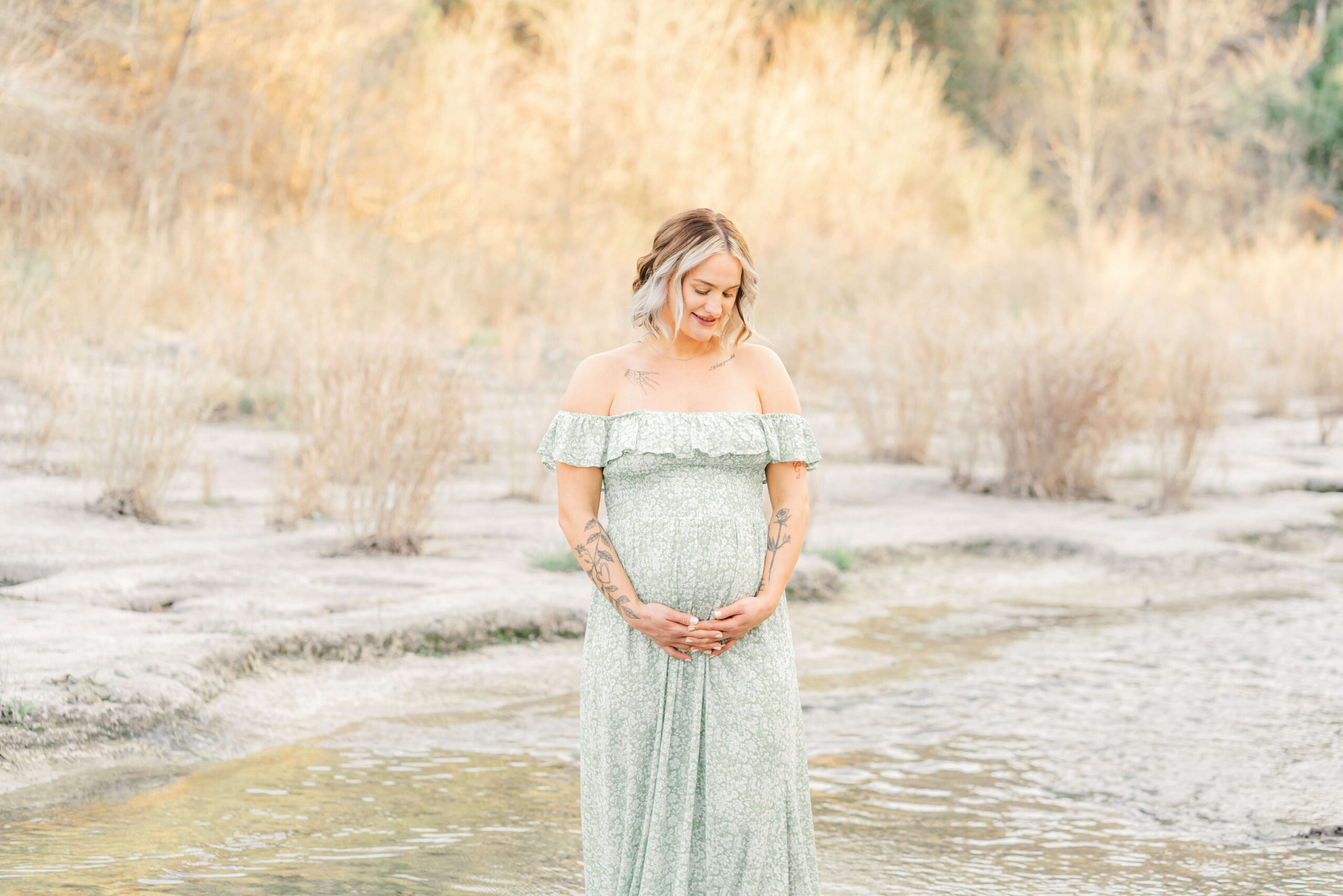 A smiling pregnant woman gazes down to her bump while standing in a creek in a green maternity dress