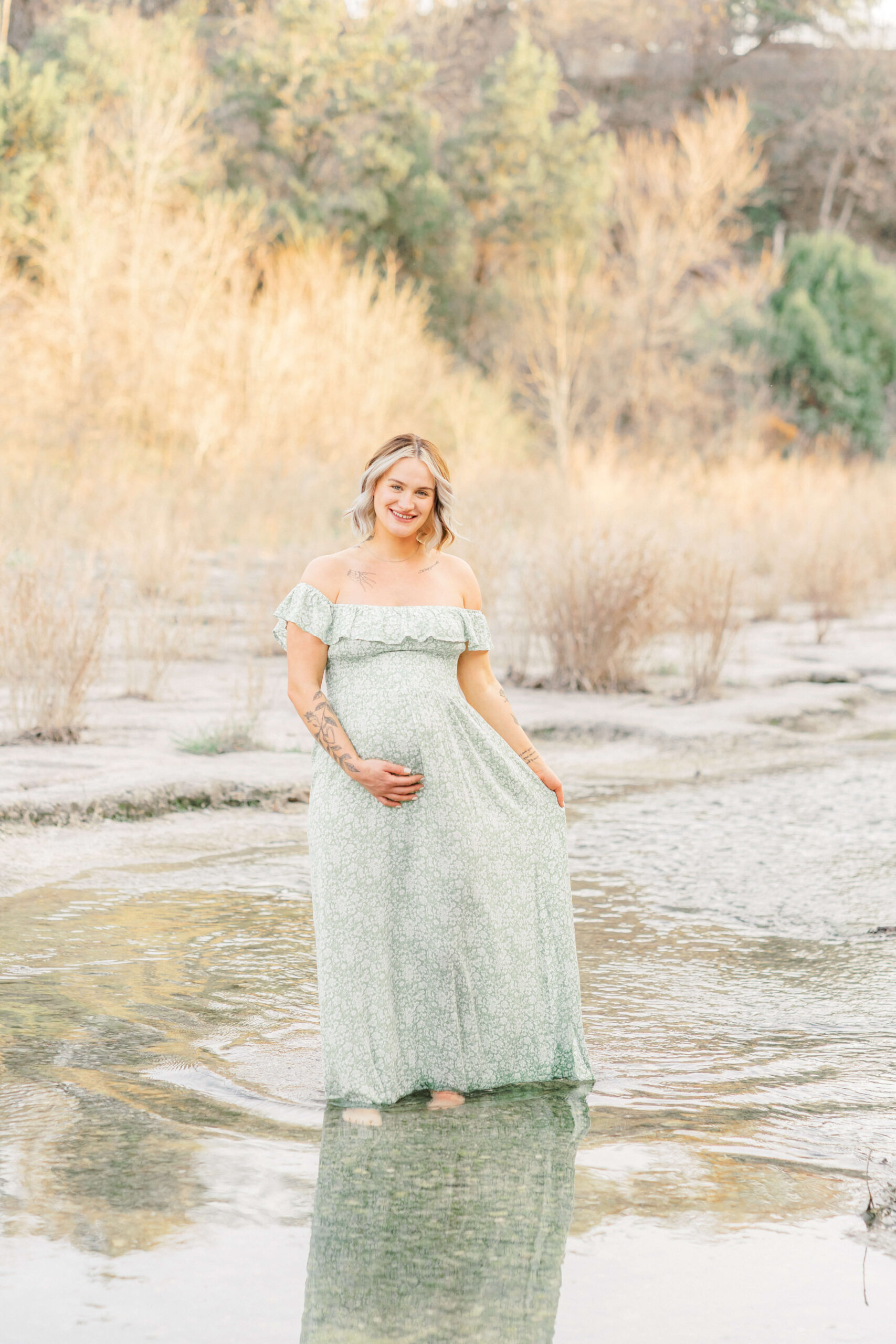 A mom to be walks in a shallow creek in a green maternity gown playing with her train after a 3d ultrasound in austin