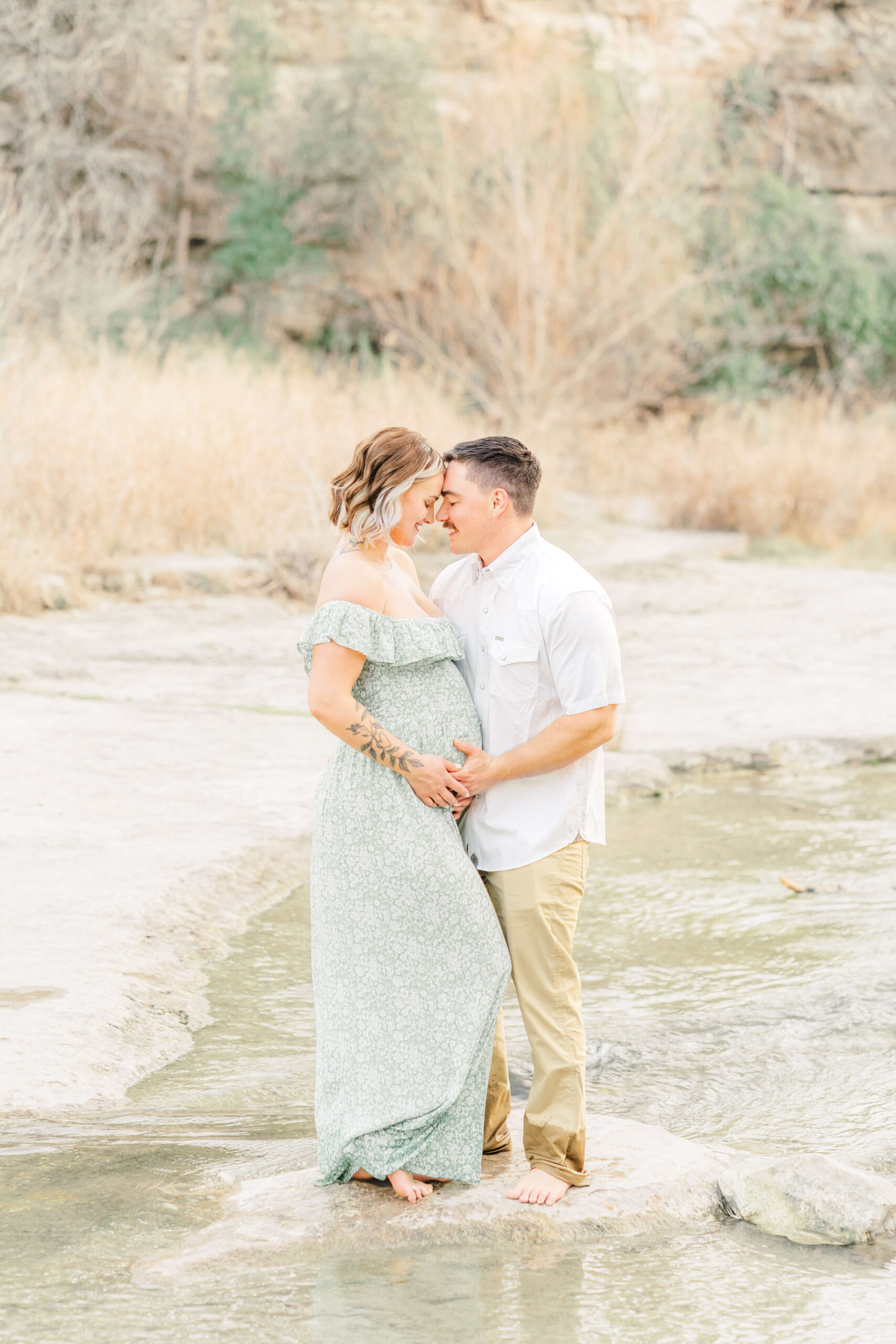An expecting couple in a green dress and white shirt snuggle touching foreheads on a boulder in a creek after their 3d ultrasound in austin