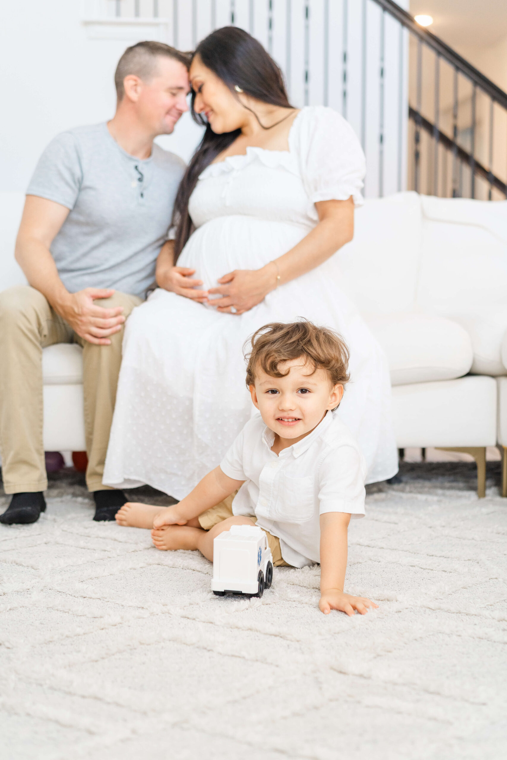 A toddler boy in a white shirt sits on the living room rug while pregnant mom and dad snuggle on the couch behind him after visiting birth centers in austin