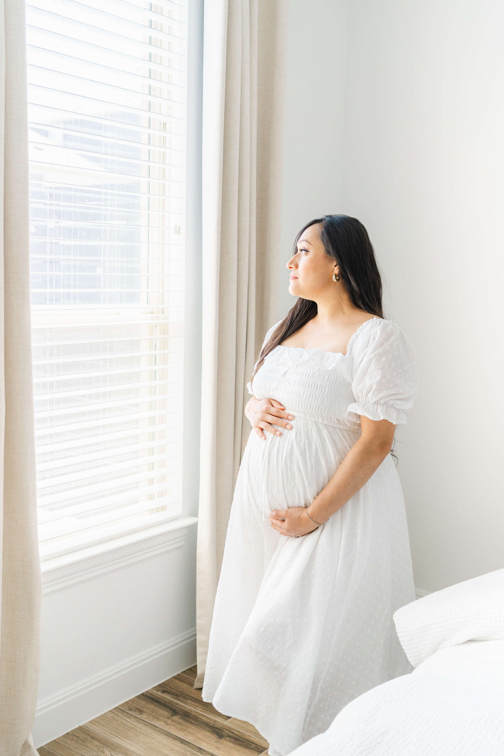 A mom to be gazes out a window while holding her bump in a studio in a white maternity gown after visiting birth centers in austin