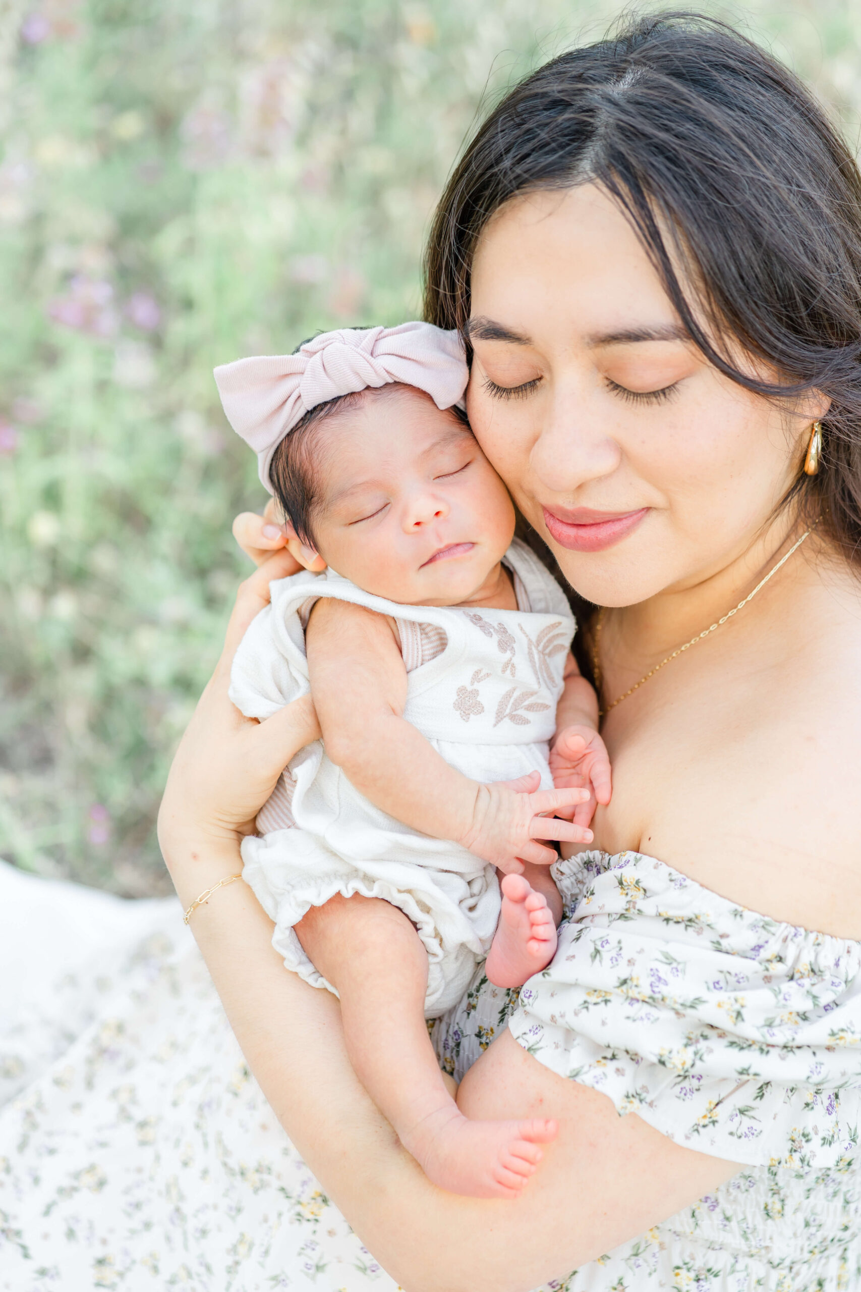 A happy new mom in a floral print dress snuggles her newborn to her cheek while sitting in a field