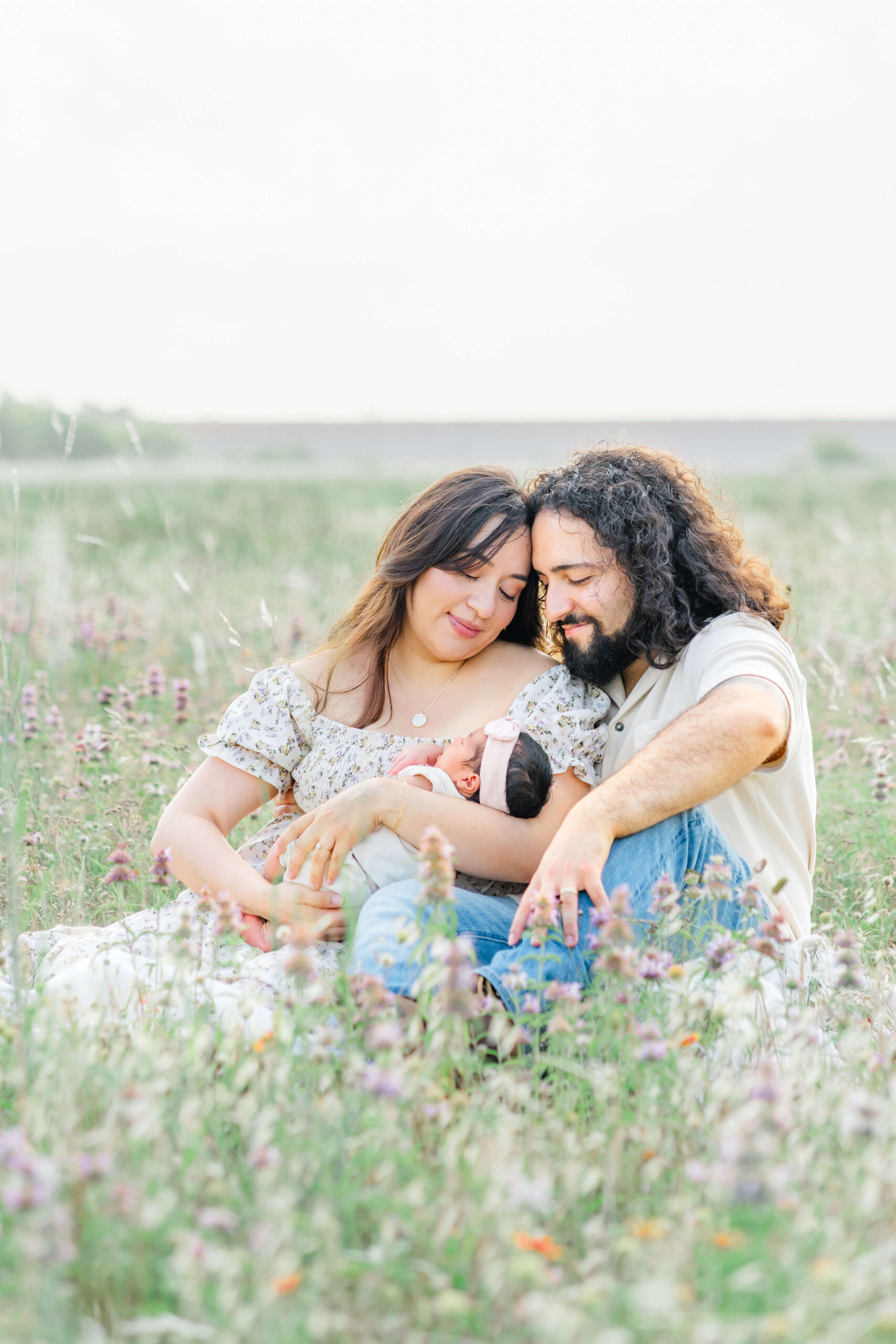 Happy new mom and dad snuggle in a field of wildflowers with their newborn sleeping in their laps after meeting lactation consultants in austin