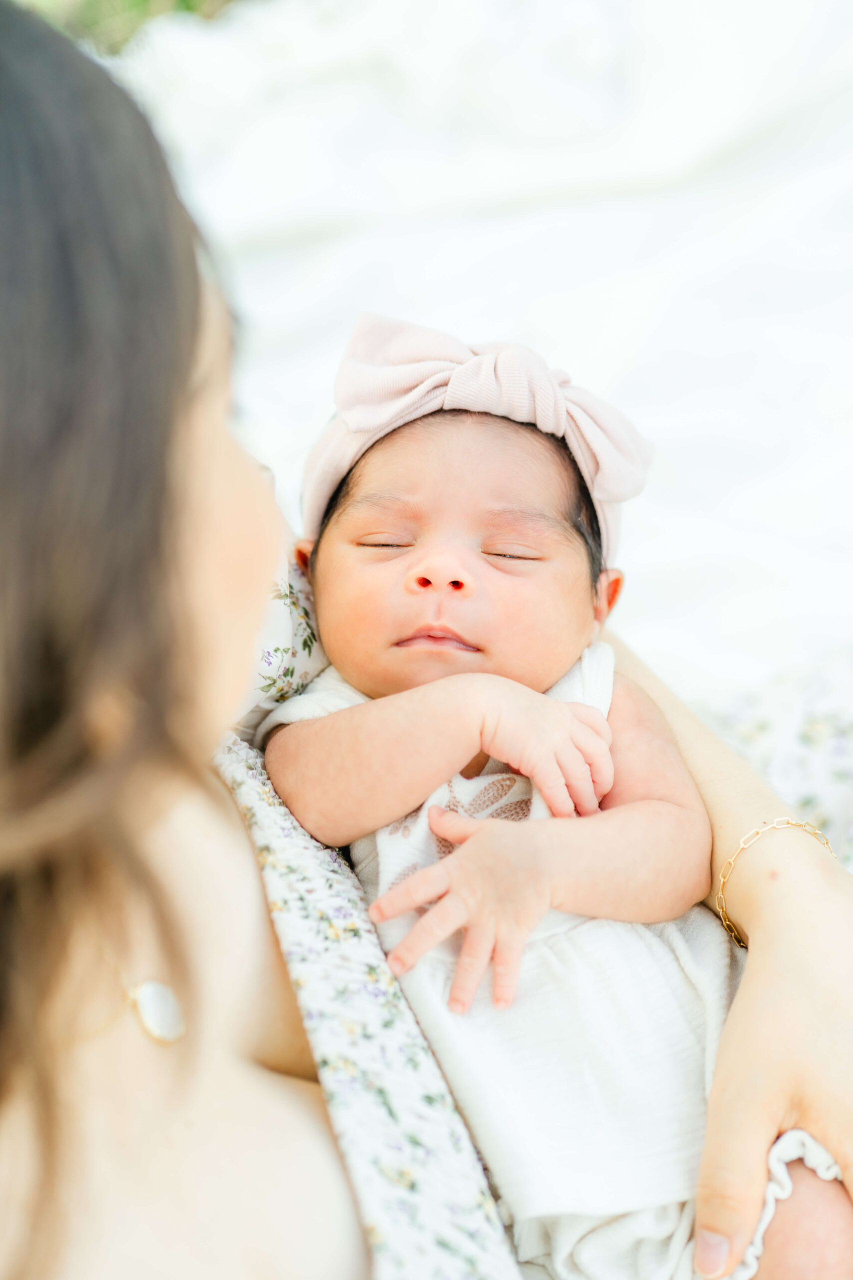 A newborn baby in a cream bow sleeps in mom's arms in a white dress thanks to lactation consultants in austin
