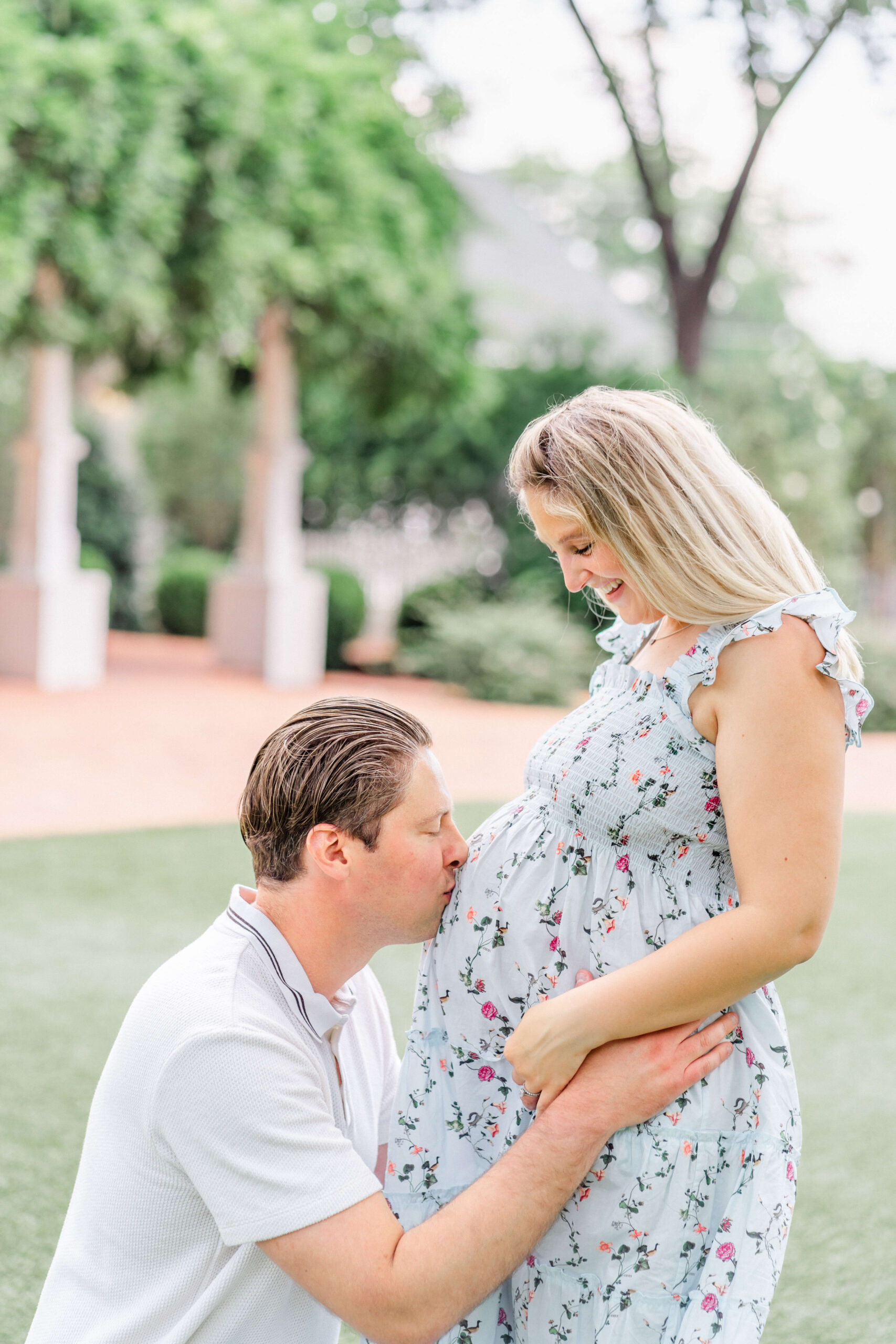 A man kisses the pregnant bump of his wife while kneeling in a park as she wers a blue floral print dress after meeting midwives in austin