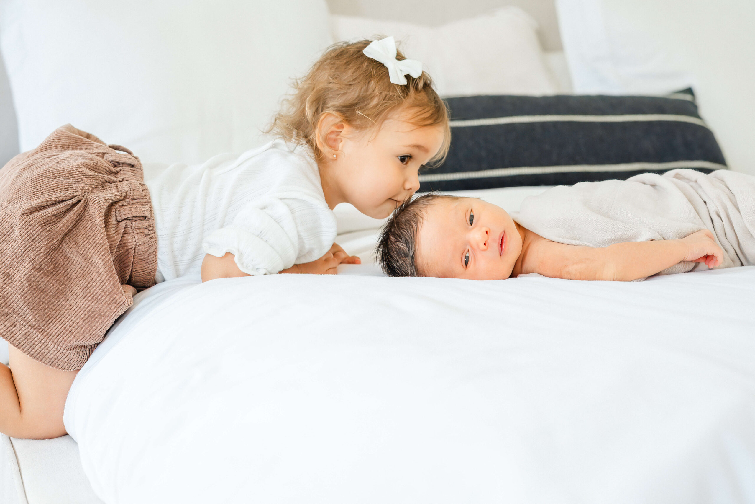 A toddler girl in a white shirt kisses the head of her newborn baby sibling laying under a blanket on a bed